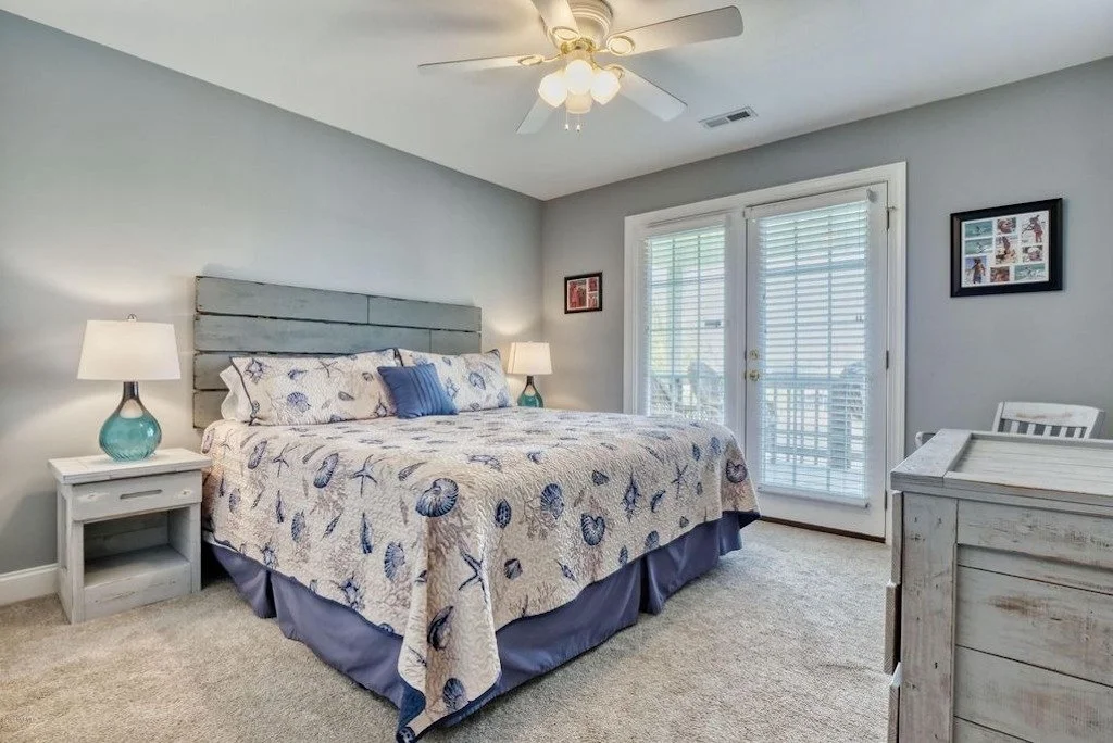 A bedroom with a bed dressed in marine-themed bedding, two nightstands with lamps, a ceiling fan, and a sliding glass door leading to a balcony.