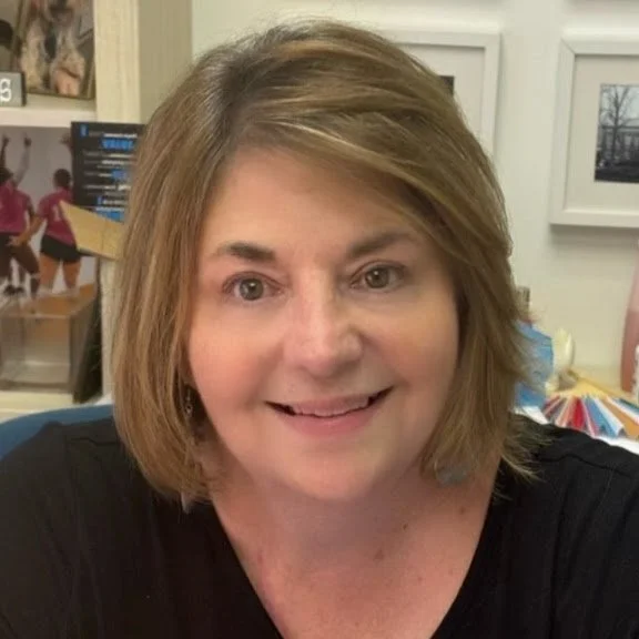 Close-up of a woman with light brown, shoulder-length hair smiling at the camera, in an indoor setting with framed pictures and colorful items in the background.