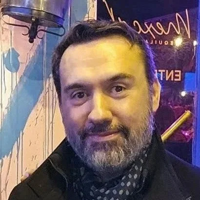 A man with dark hair and a beard, smiling in front of colorful neon lighting.