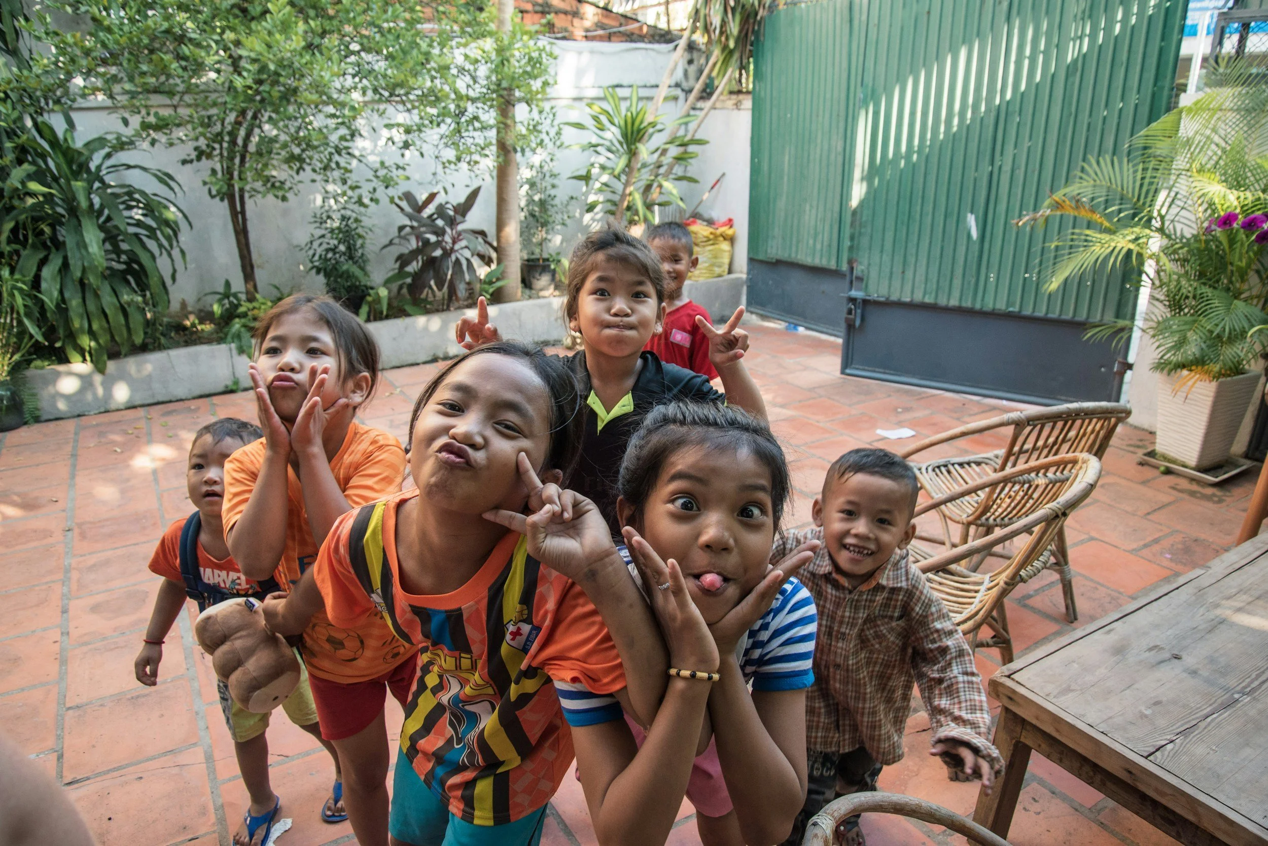 A group of children making funny faces and posing for a photo outdoors on a brick patio surrounded by greenery, with a green metal gate and potted plants in the background.