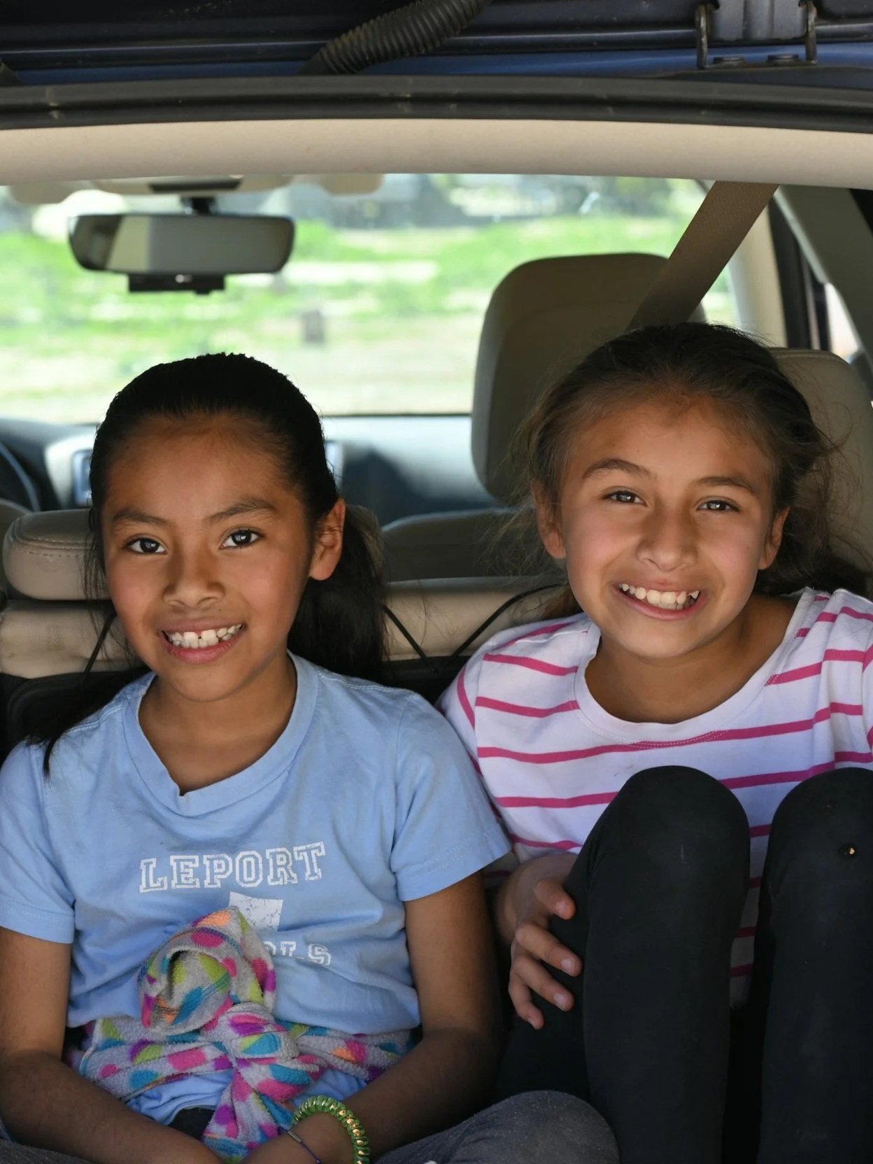 Two young girls sitting in the backseat of a car, smiling at the camera. One has dark hair in pigtails, wearing a light blue shirt, and the other has lighter hair, wearing a pink and white striped shirt.