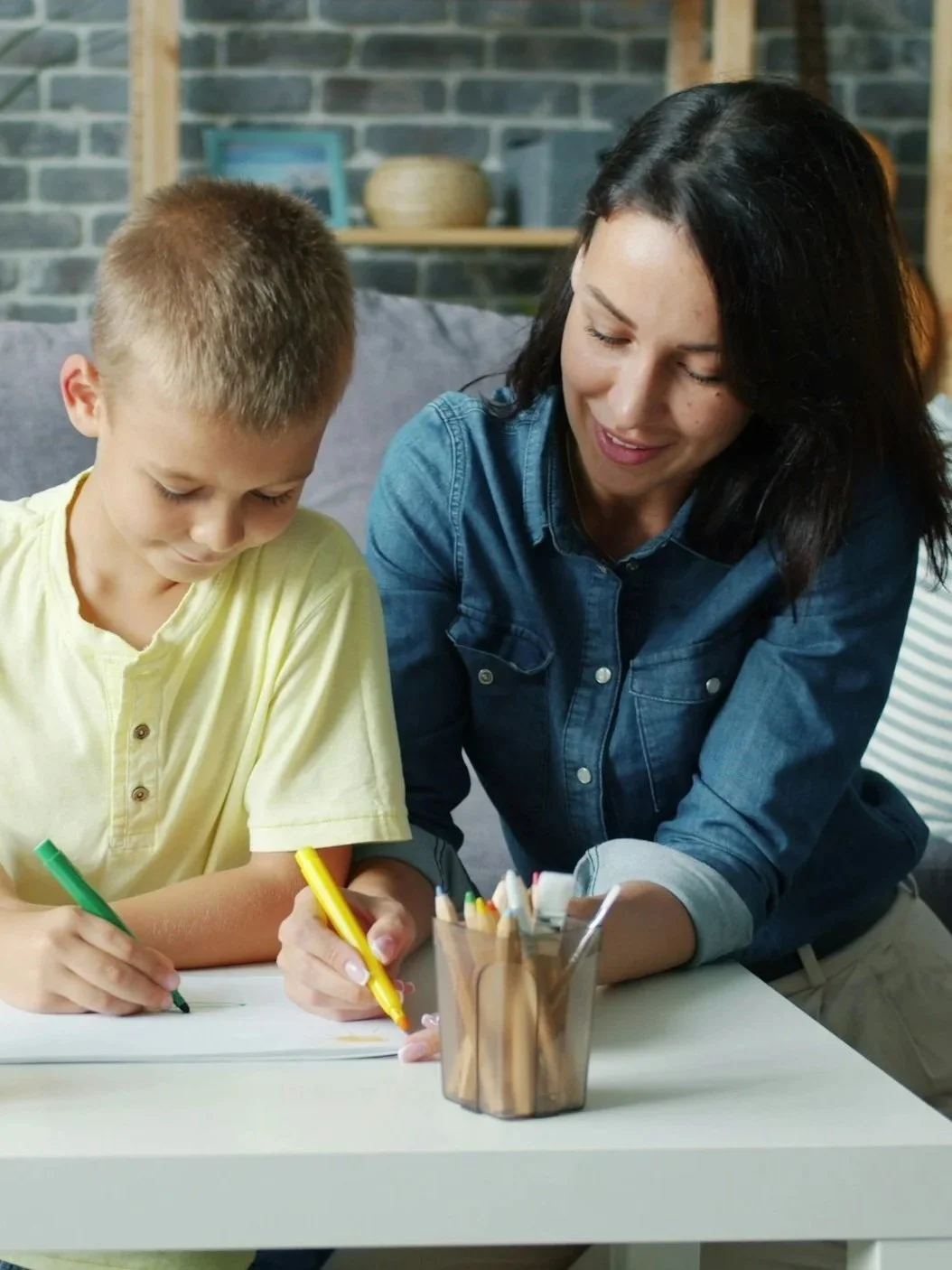 A woman and a boy drawing with colored markers at a table in a cozy room with a brick wall background.