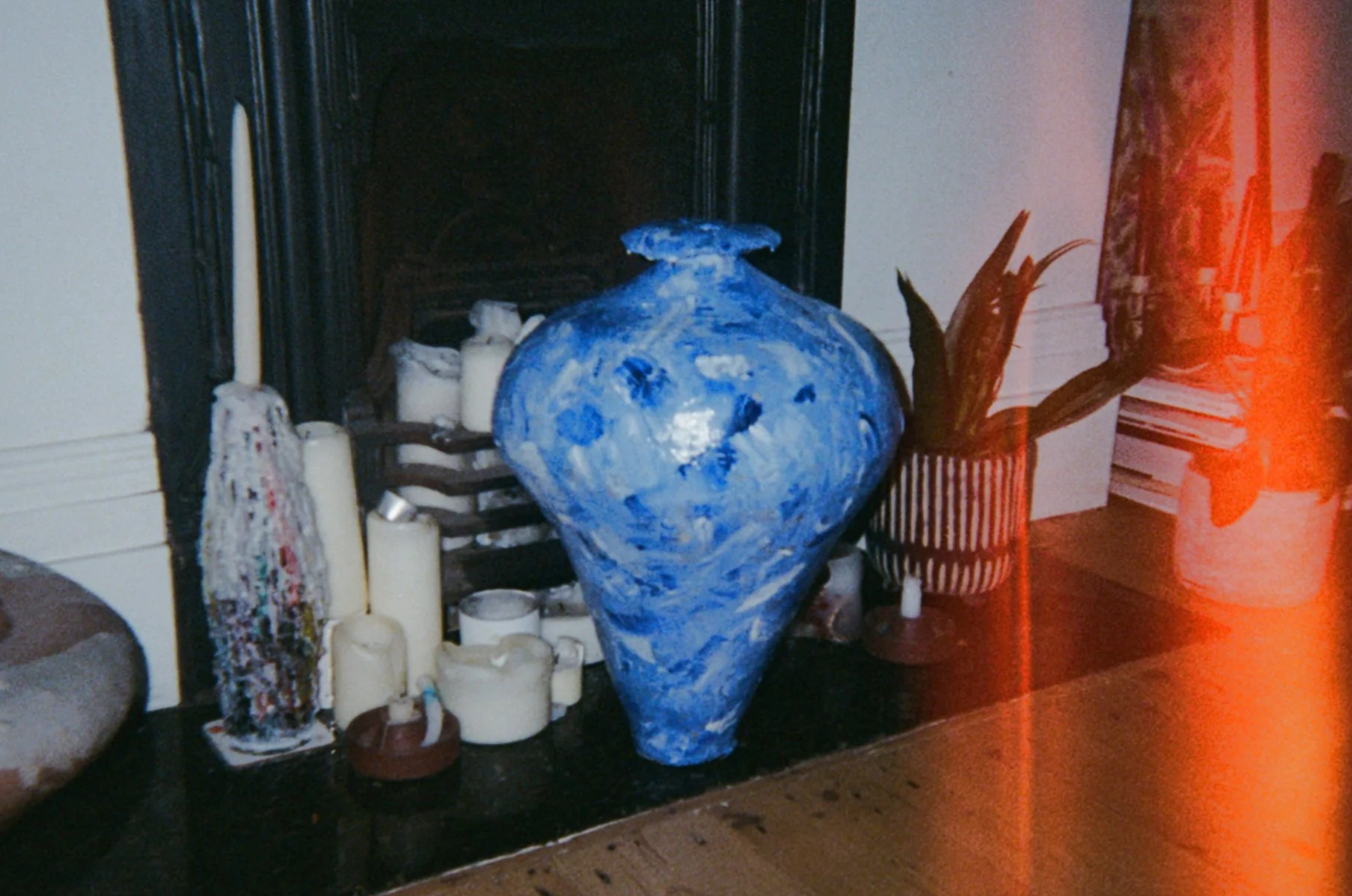 A large blue and white ceramic vase surrounded by numerous candles of various sizes and a patterned fabric. There is also a plant in a striped pot against a white wall, with a wooden floor visible at the bottom. A section of a black fireplace backdrop is visible behind the objects.
