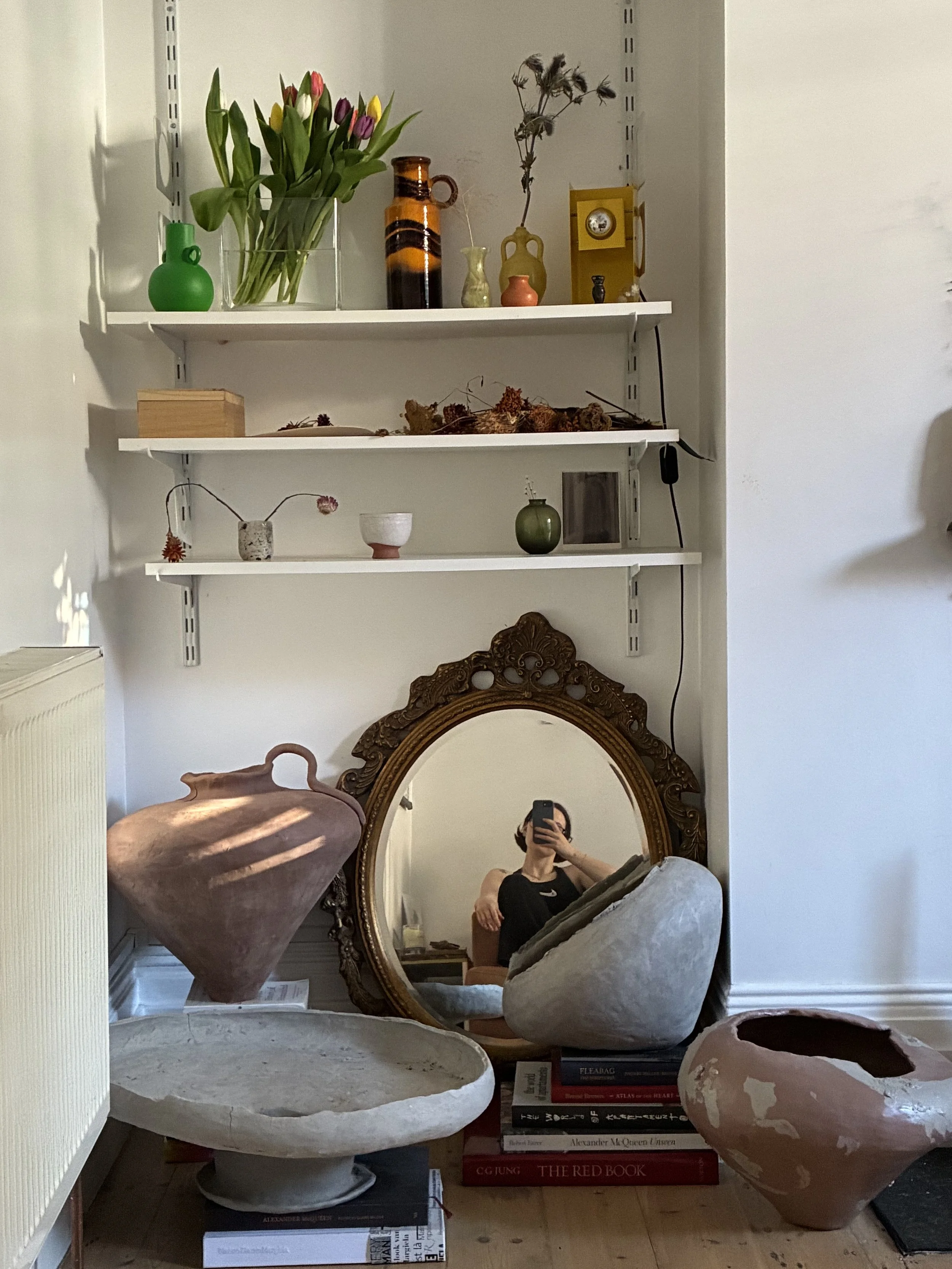 A corner of a room with three white shelves filled with decorative items including vases, books, and dried flowers. A large ornate mirror leans against the wall, reflecting a person taking the photo. The room has light-colored walls and a wooden floor.