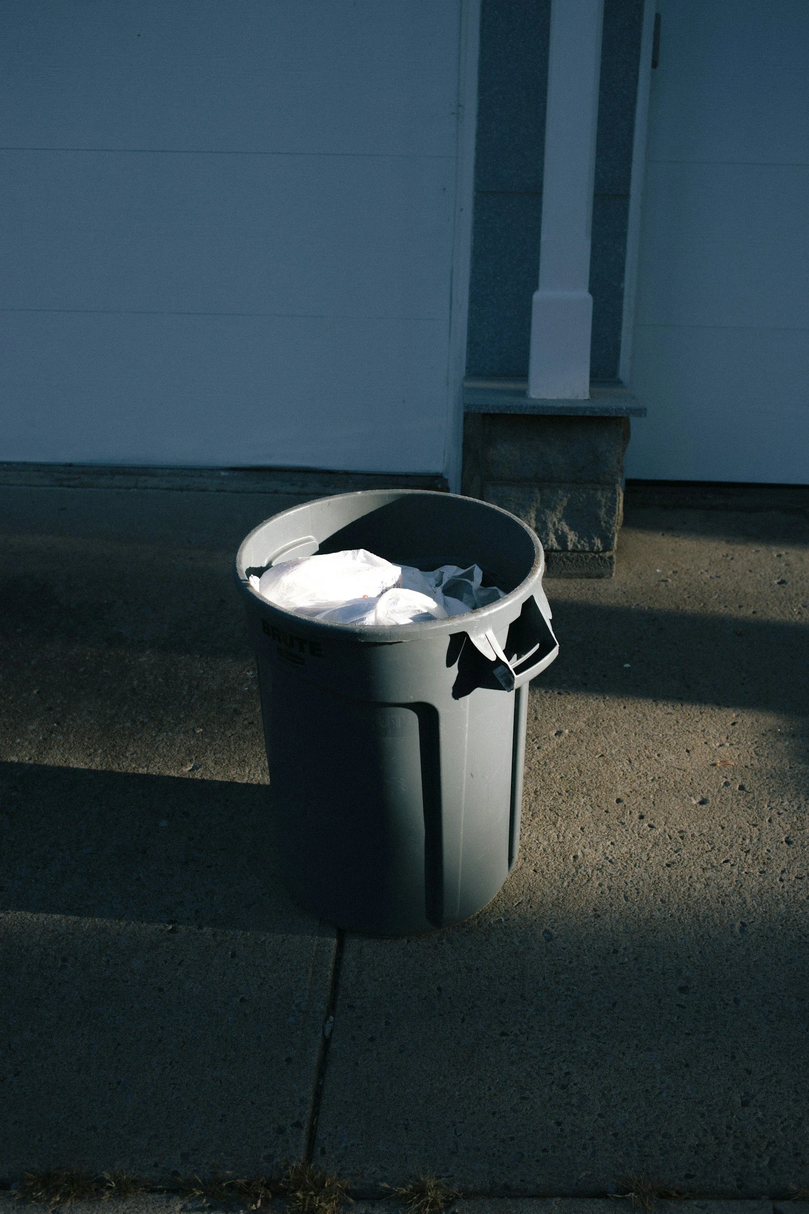 A white trash can on the street with trash inside, in front of a building with a white wall, a door, and a small window.
