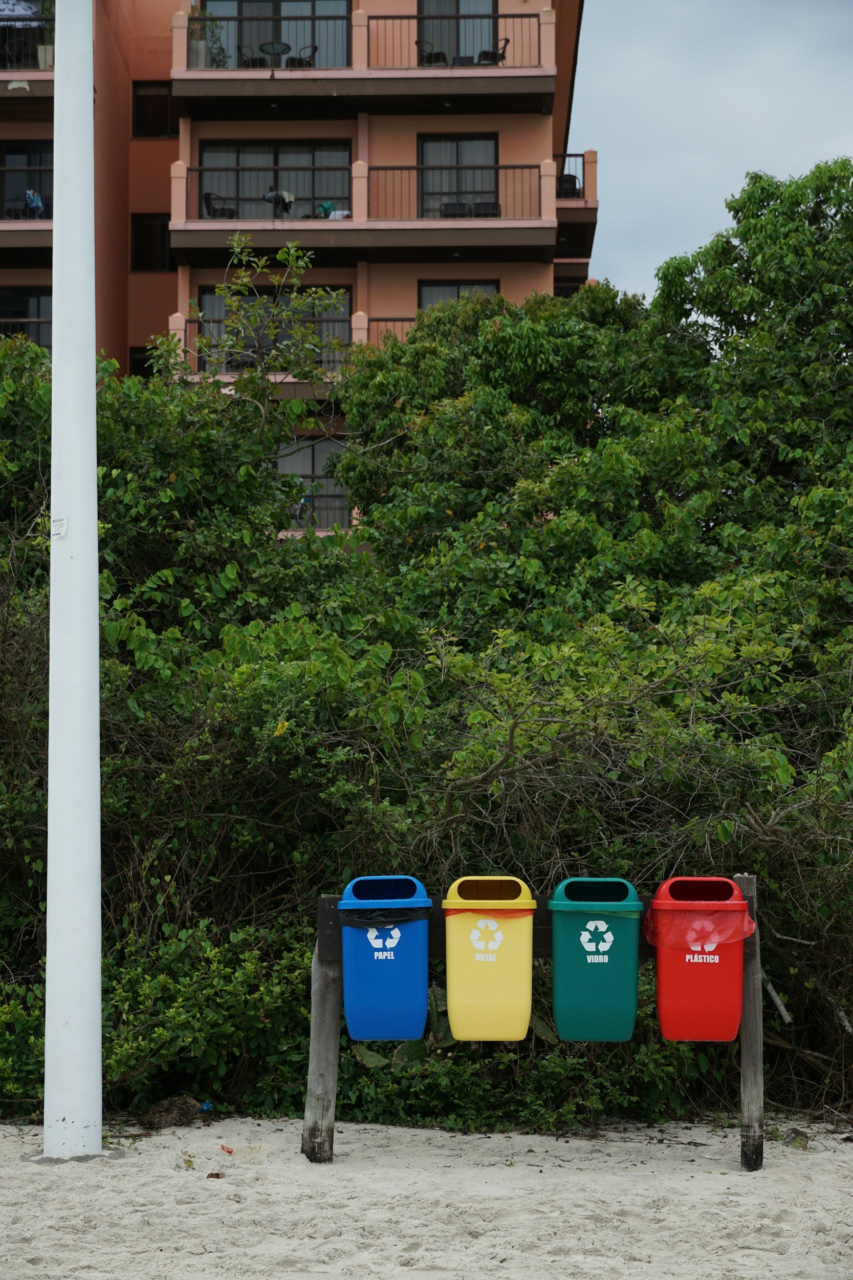 Four trash bins in blue, yellow, green, and red colors labeled for recycling categories—paper, metal, glass, and plastic—are placed on a sandy surface in front of a green hedge and a building with balconies.