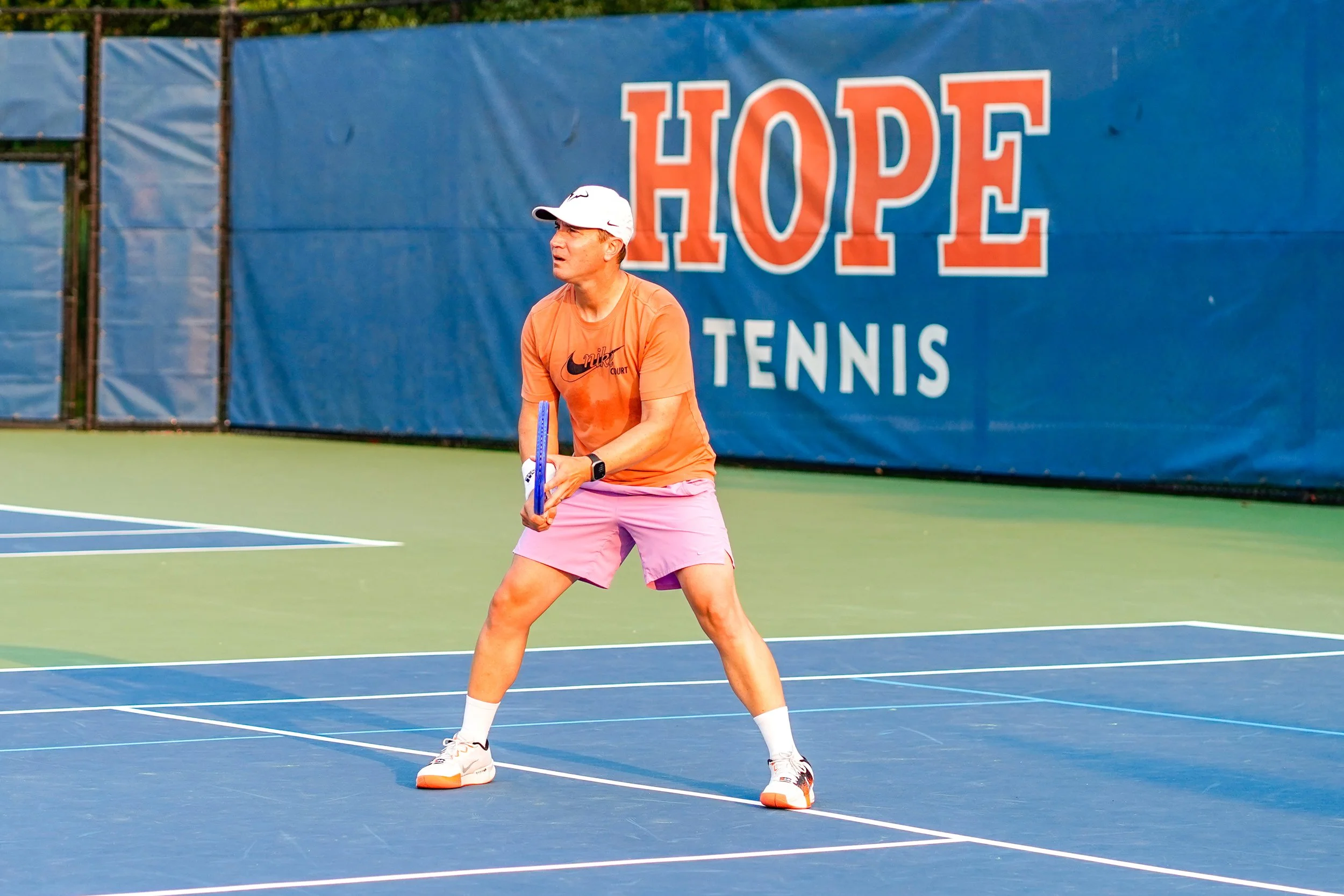 A man in an orange Nike shirt and pink shorts is playing tennis on an outdoor court. He is holding a tennis racket with both hands, standing in a ready position, and wearing a white cap and white/black sports shoes. Behind him is a blue backdrop with a large sign that says 'HOPE' in orange and white letters, and 'TENNIS' in white letters.
