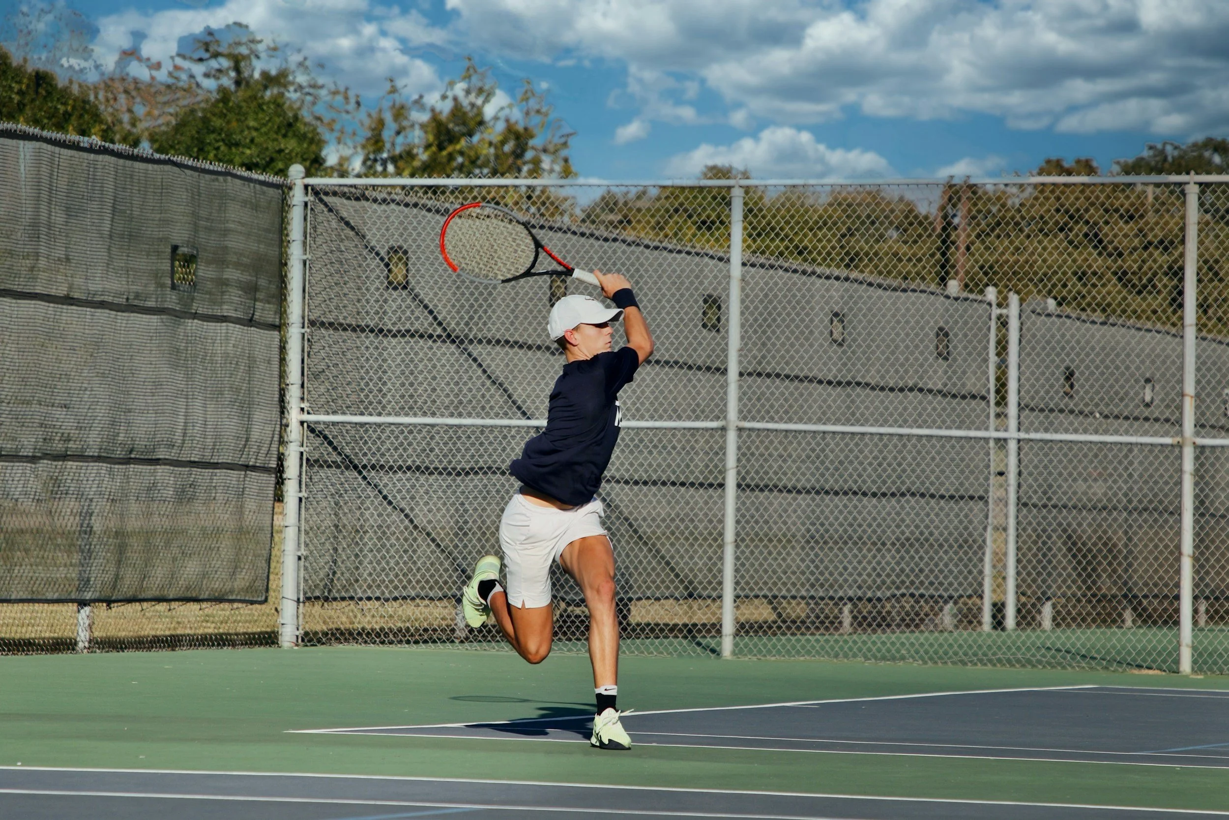 A young man hitting a tennis ball with a racket on an outdoor tennis court, wearing a white cap, black shirt, white shorts, and tennis shoes, with a chain-link fence and trees in the background under a partly cloudy sky.