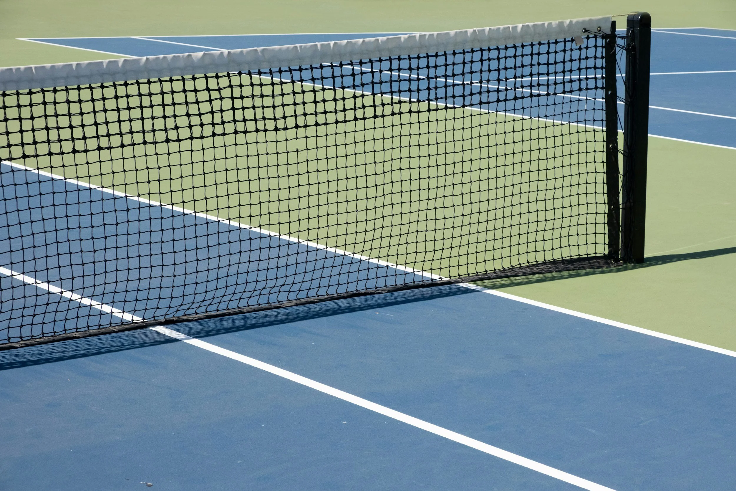 An outdoor tennis court with a black net in the foreground, white boundary lines, and a green and blue surface.