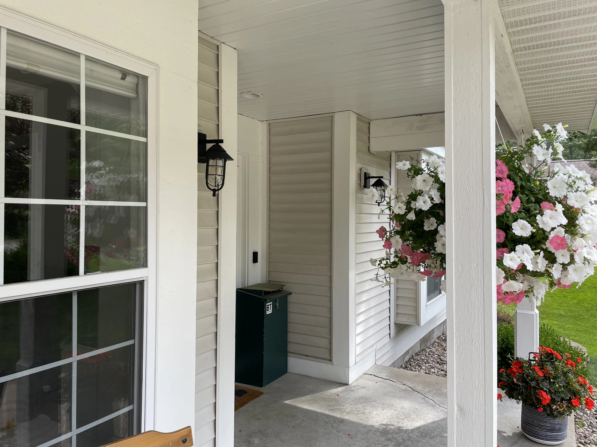 Front porch area of a house with a large window, black outdoor wall lanterns, and a white porch support column with potted flowers, including red and white petunias.
