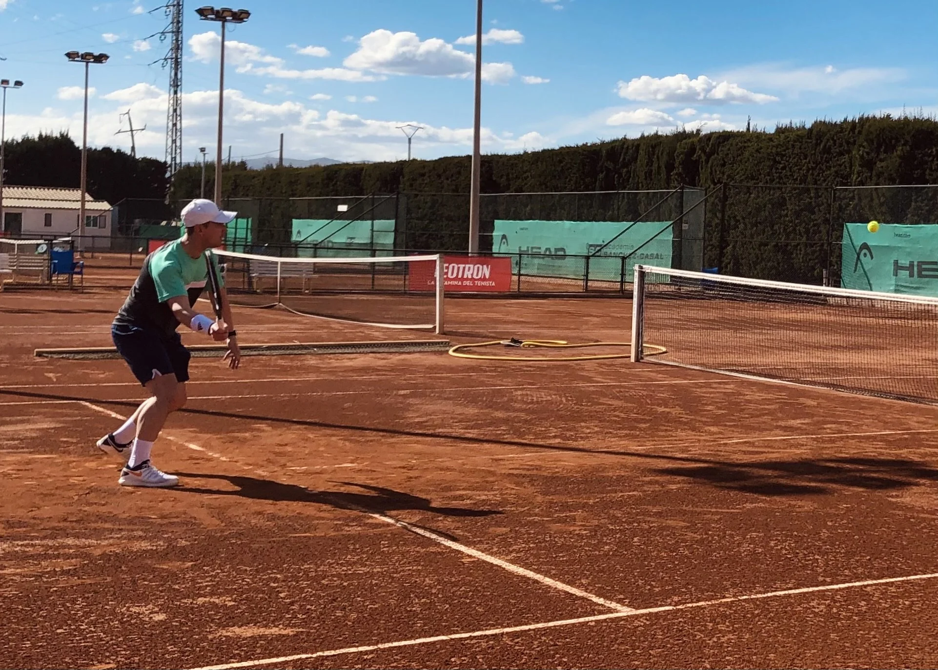 A man playing tennis on a clay court, preparing to hit a tennis ball, with the court's net in the foreground and a fence, banners, and trees in the background under a partly cloudy sky.