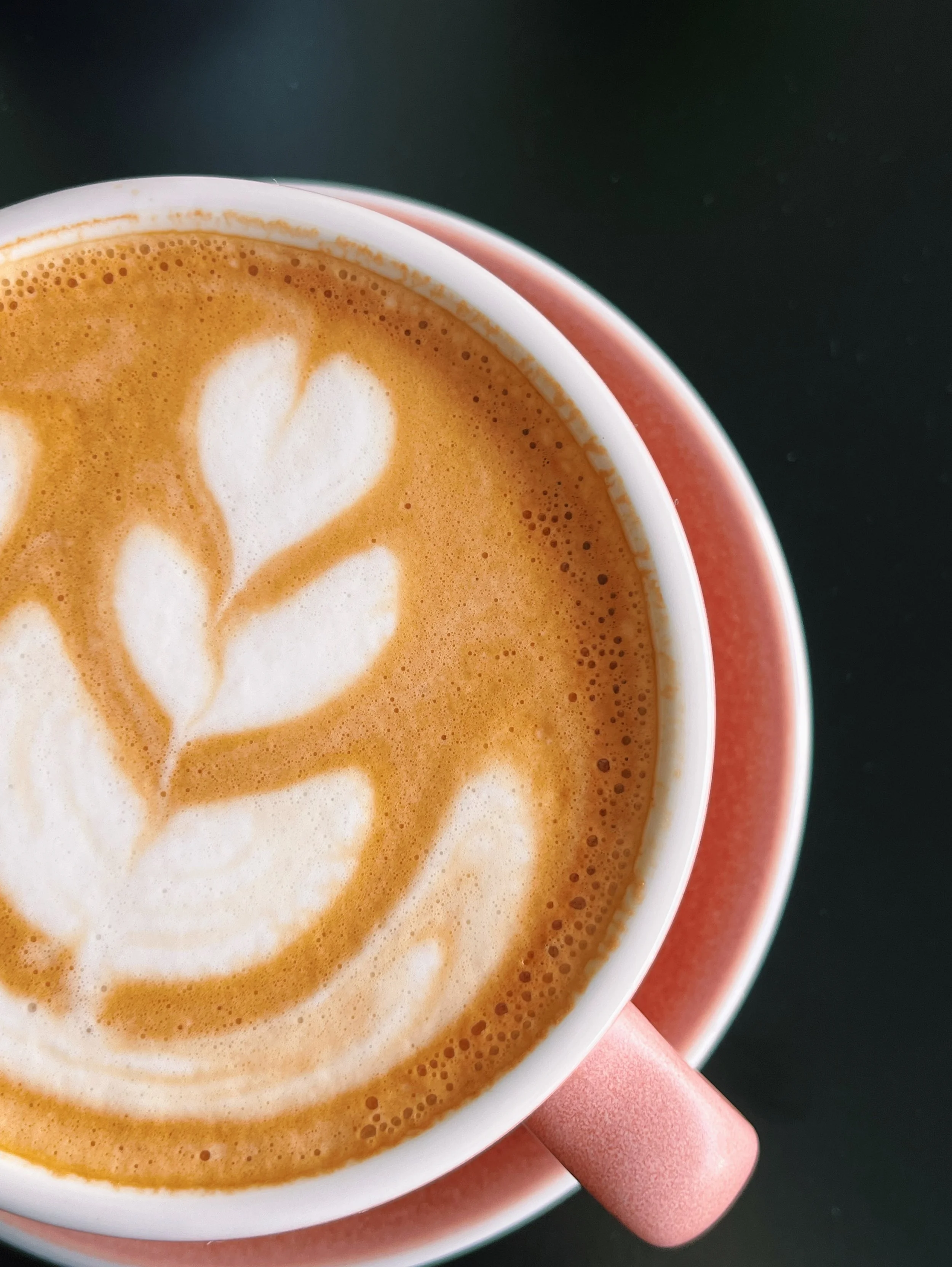 A close-up of a cup of coffee with latte art in the shape of hearts, held in a pink-handled mug on a dark surface.