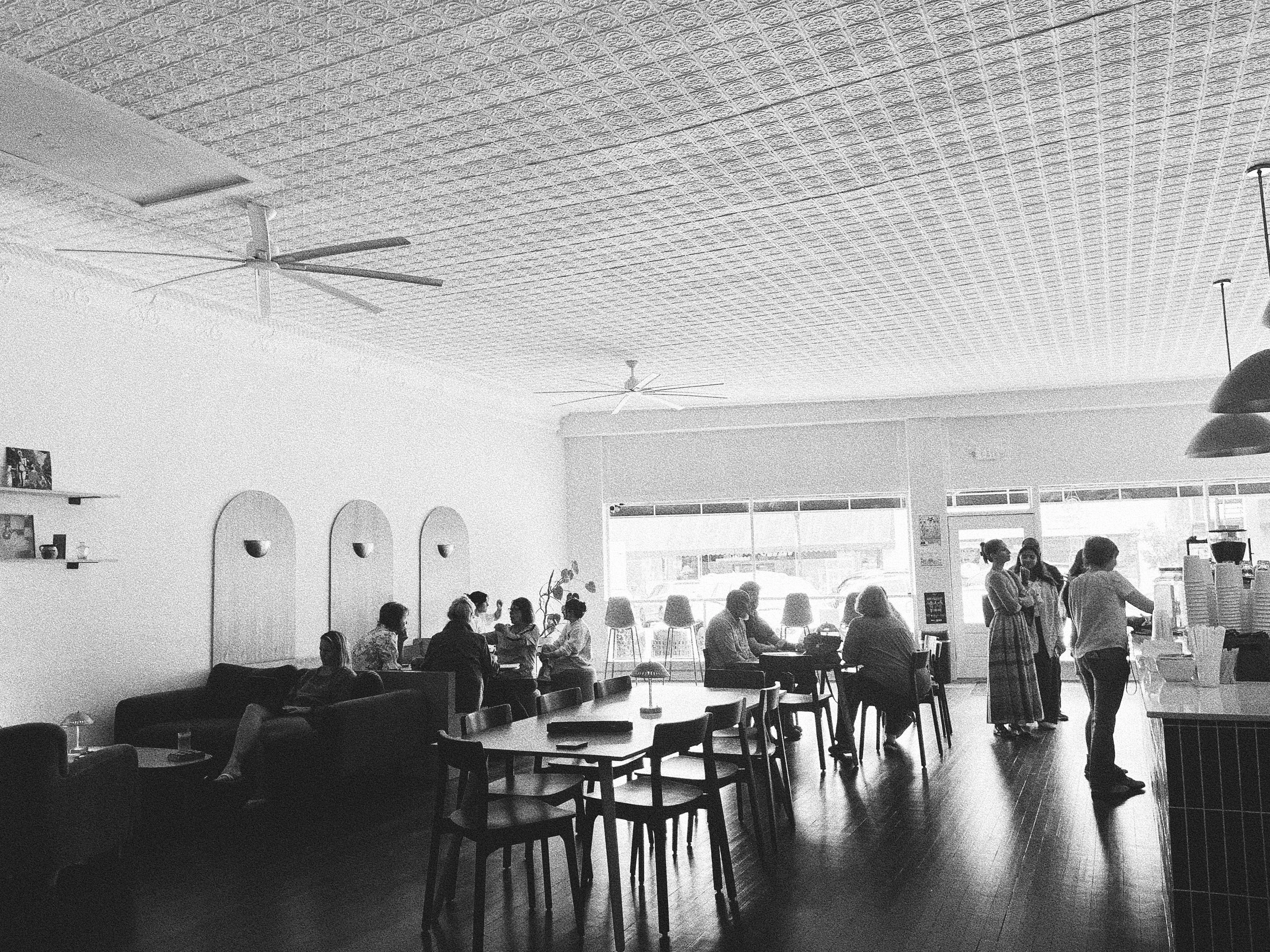 Interior of a cozy cafe with people sitting at tables, some chatting and others using their phones. The cafe features a textured ceiling, ceiling fans, and large windows allowing natural light to brighten the space.