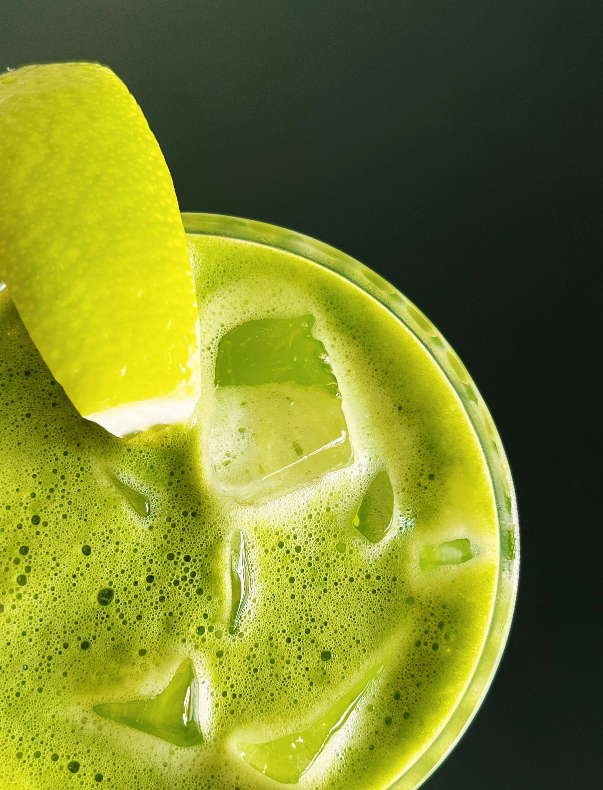 Close-up of a glass of bright green matcha latte with ice cubes, topped with a lemon wedge, against a dark background.