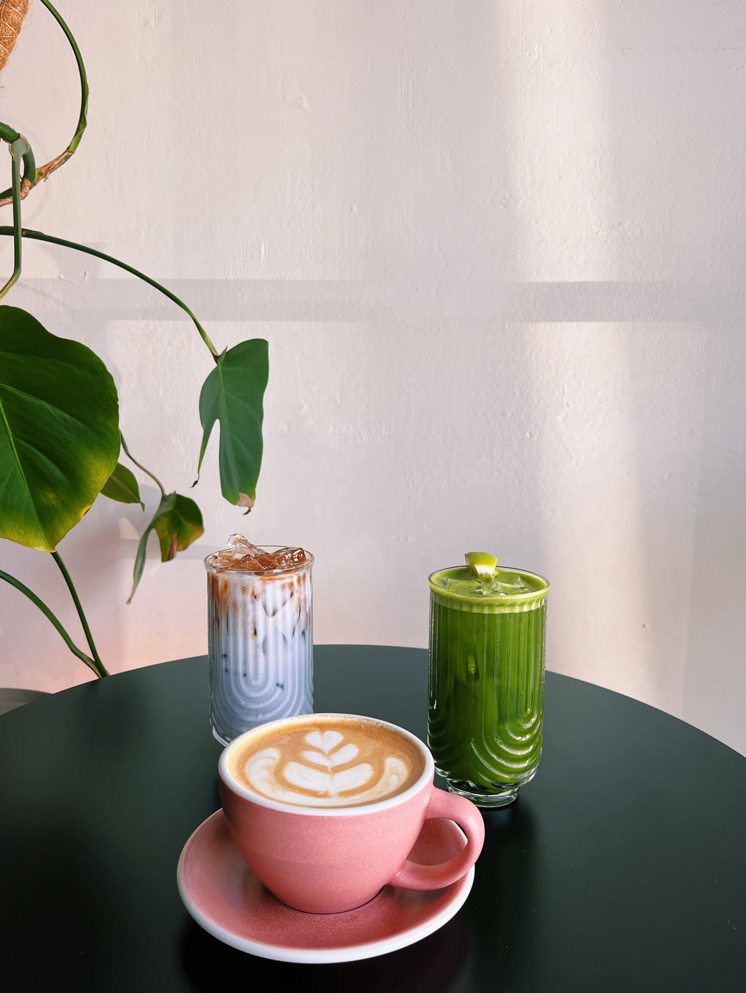 A pink ceramic cup with a matching saucer on a black table, filled with a latte art coffee featuring a tulip pattern, two glasses of iced beverages (one with a creamy appearance and the other green), and a green houseplant with large leaves against a white wall.