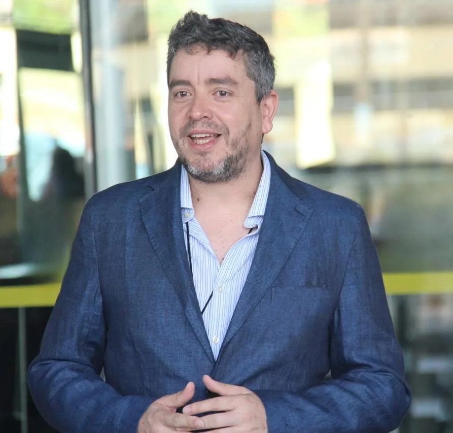 Homem sorridente com barba, cabelos curtos e grisalhos, usando terno azul e camisa de botão azul claro, em ambiente urbano