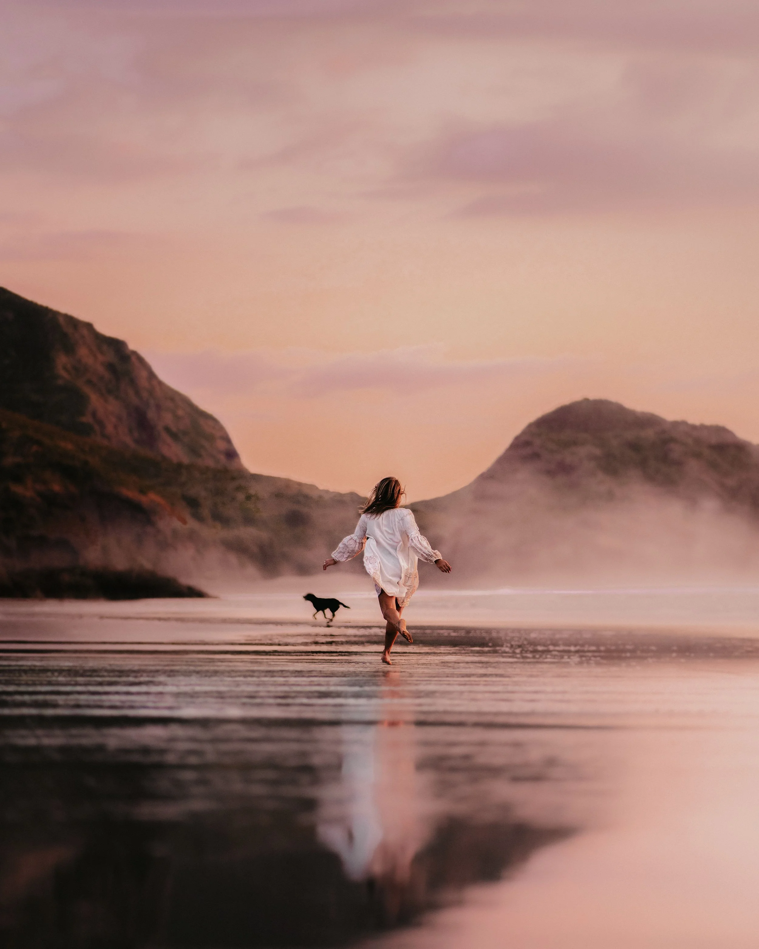 A woman in a white dress running on a beach at sunset with a dog in the background, and mountains in the distance.