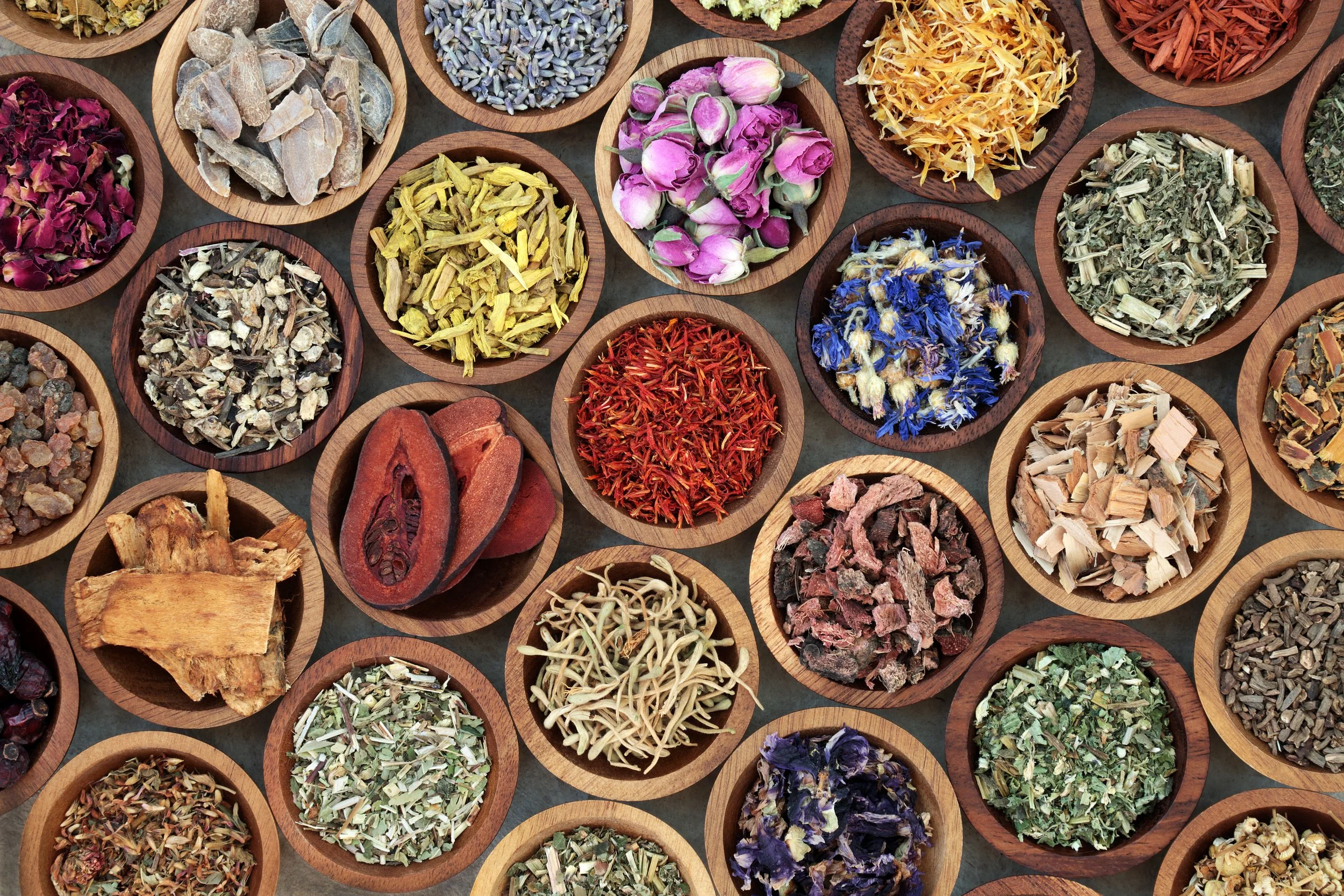 Multiple small wooden bowls containing various dried herbs, flowers, and botanicals arranged on a table.