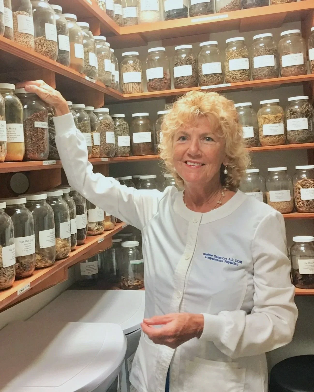 A smiling woman in a white medical coat browsing spices stored in glass jars on shelves in a apothecary or spice shop.