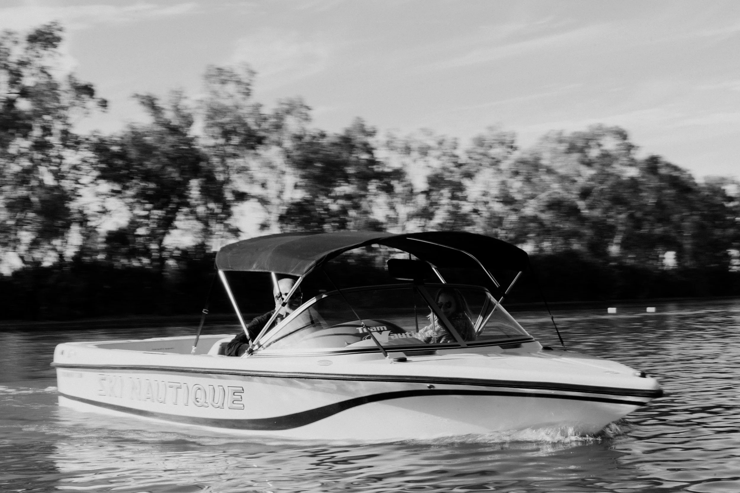A black and white photo of a small motorboat on a body of water with trees in the background. The boat has a canopy with the word 'NAUTIQUE' on the side and appears to be moving at a steady pace.