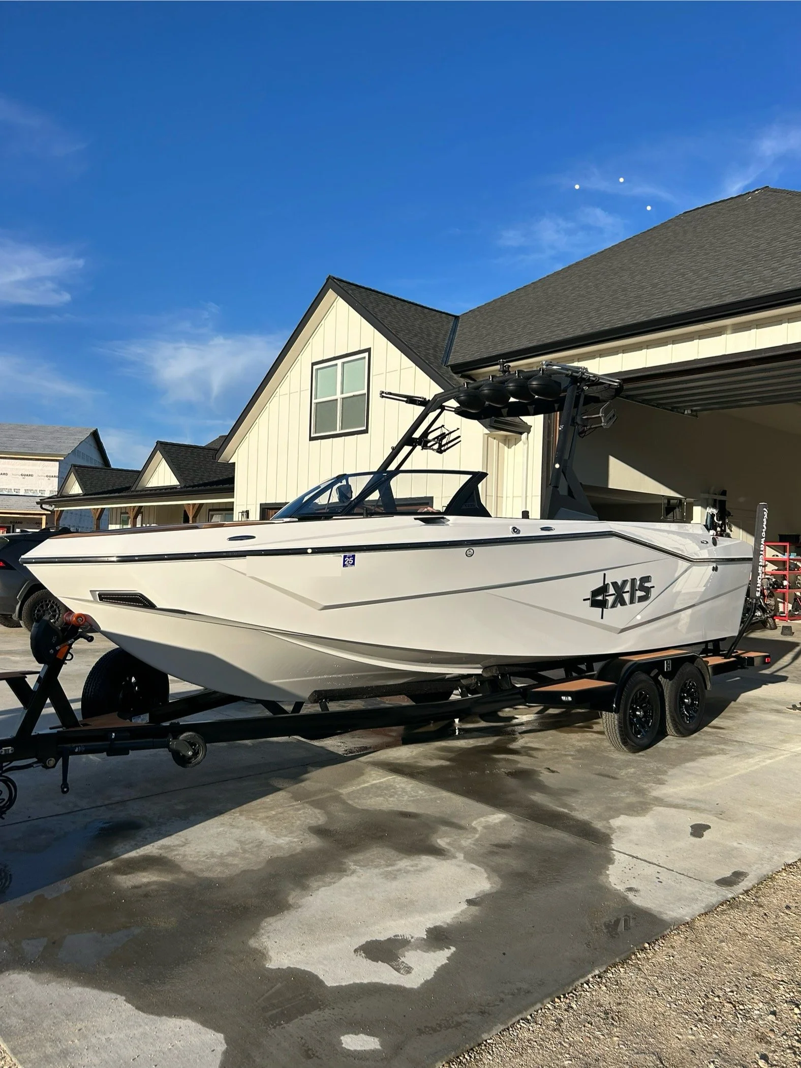 A white speedboat with black accents on a trailer in a driveway, with a house with a pitched roof and beige siding in the background, and a clear blue sky overhead.