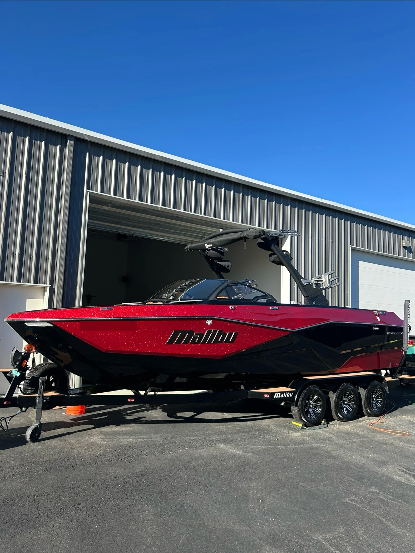 A red Malibu speedboat on a black trailer parked outdoors in front of a large metal building with a blue sky overhead.
