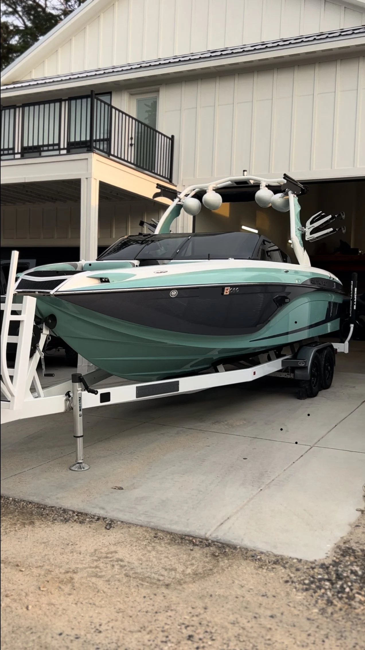 A green and black speedboat on a white trailer parked outside a house with a white exterior, a balcony with a black railing, and a garage underneath.