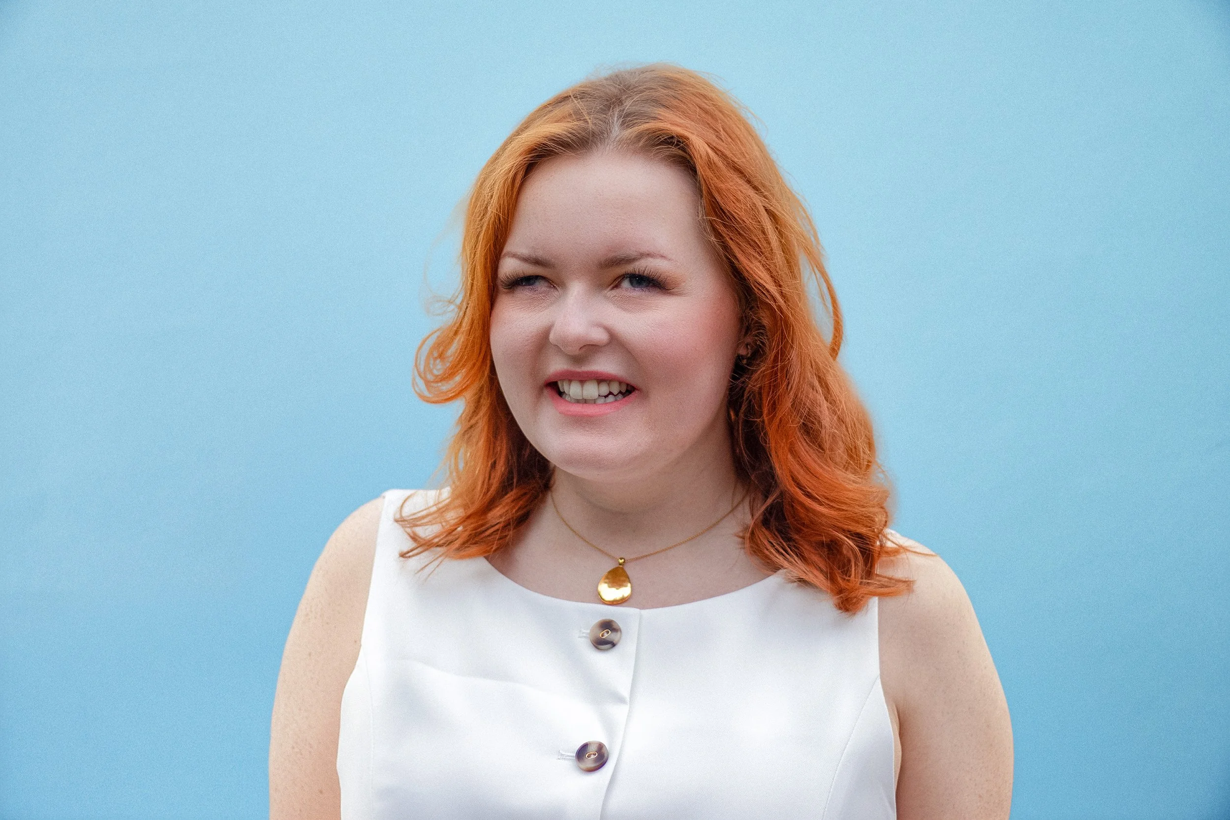 Lucy, a woman with red hair, blue eyes, and a playful expression, wears a white sleeveless top with large decorative buttons, a gold necklace with a pendant, and a light blue background.