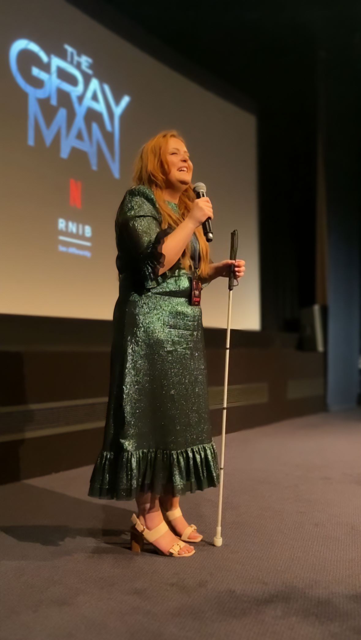 Lucy wearing a dark green, sparkly dress with ruffled hem from Vampire's Wife, holds a cane and a microphone, standing on a stage with a movie title for 'The Gray Man' projected behind her.