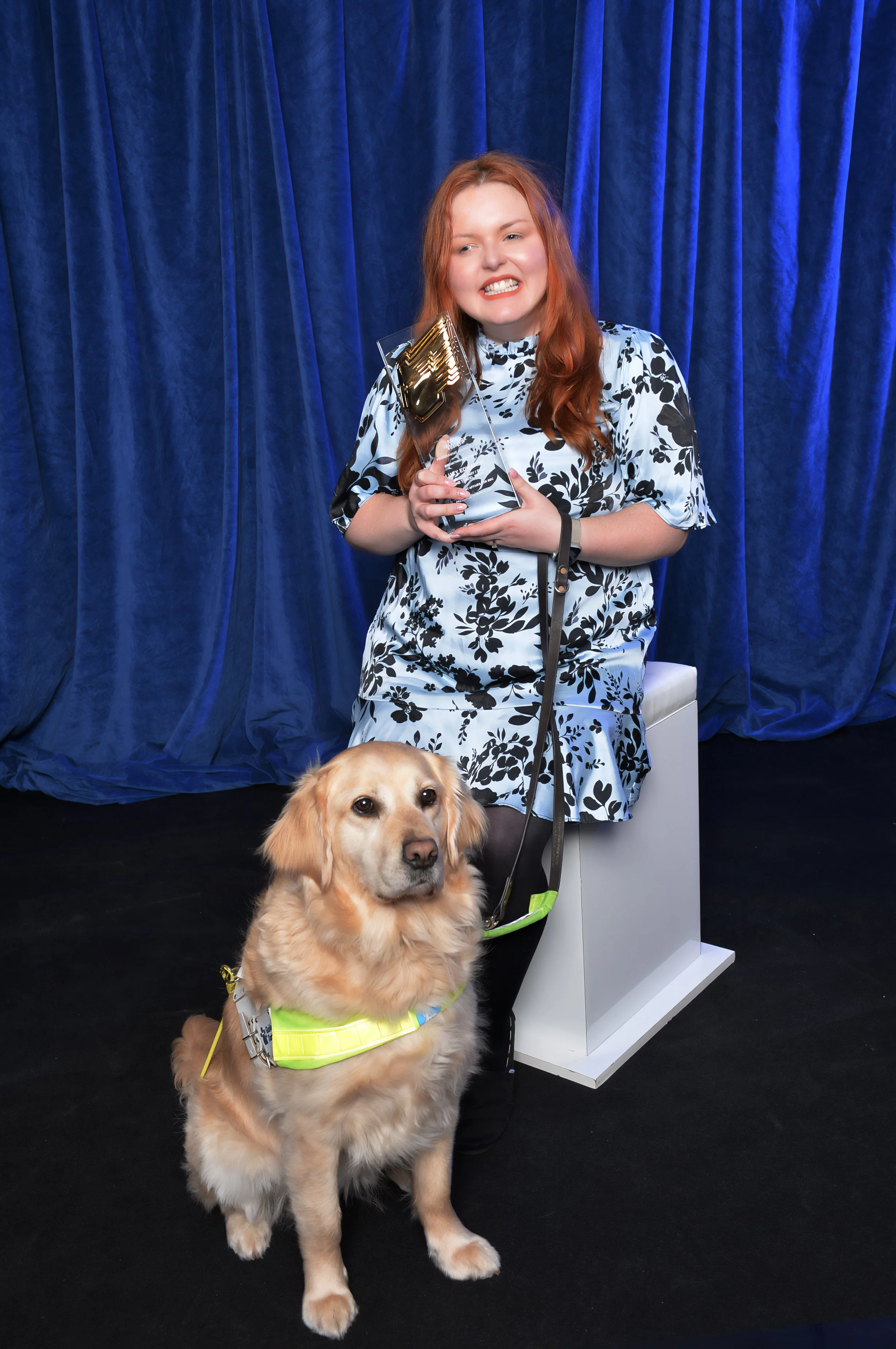 Lucy wearing a black and pale blue floral dress holds her RTS award. Guide dog Molly sits at her feet.