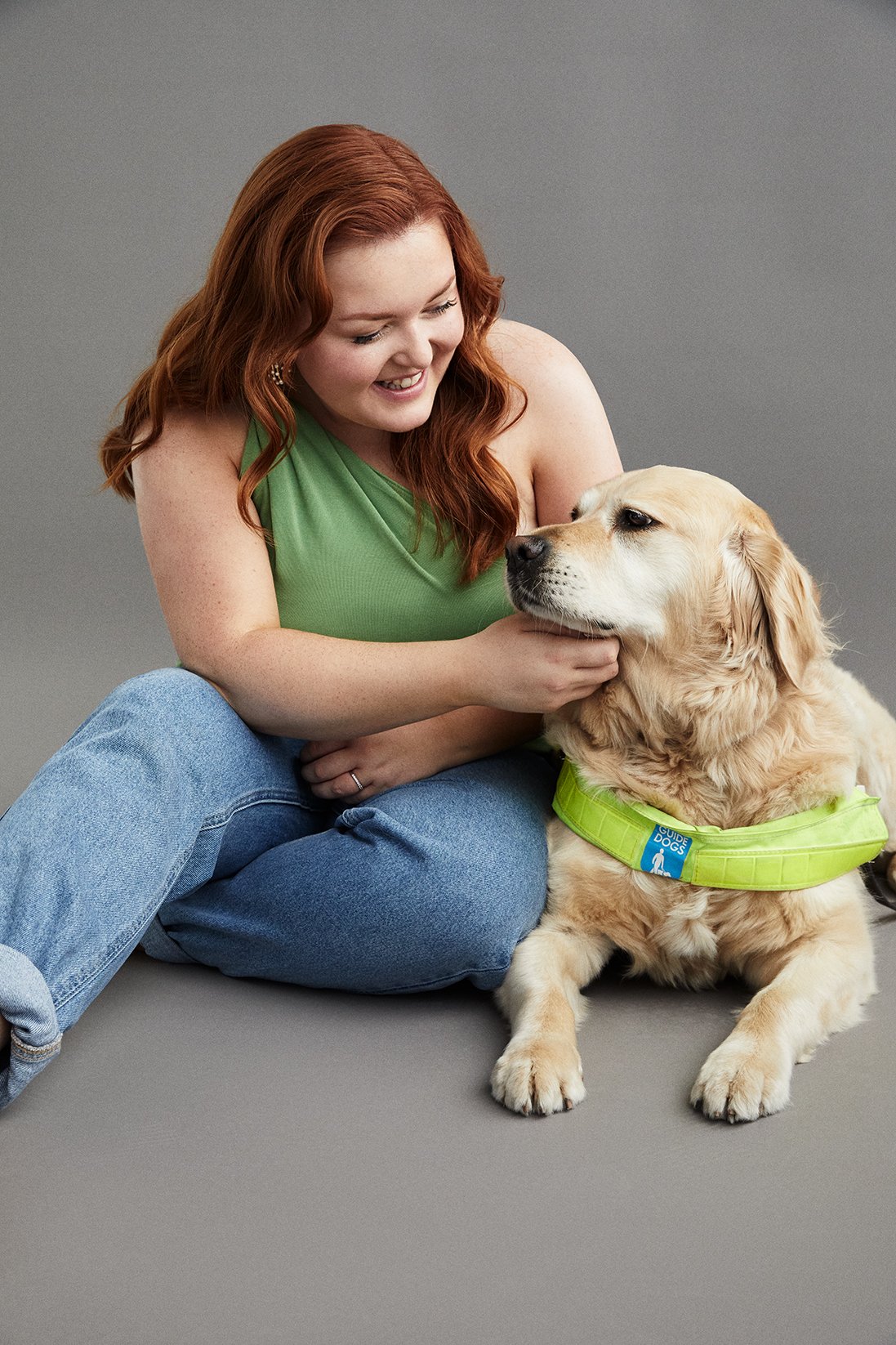 Lucy wearing a green top and blue jeans sits on the floor with  golden retriever guide dog Miss Molly who wears her guide dog harness, Lucy touches Molly's chin affectionately.