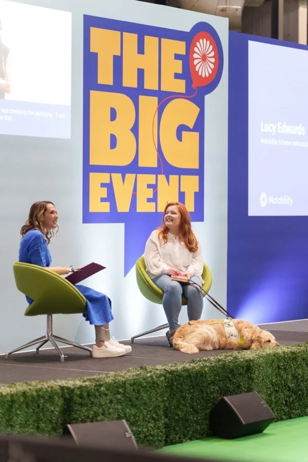 Lucy sits on a stage with a large sign reading "The Big Event" including a speech bubble and a flower icon. Her interviewer is holding a notebook, Lucy has guide dog Molly lying beside her. They are engaged in a conversation.