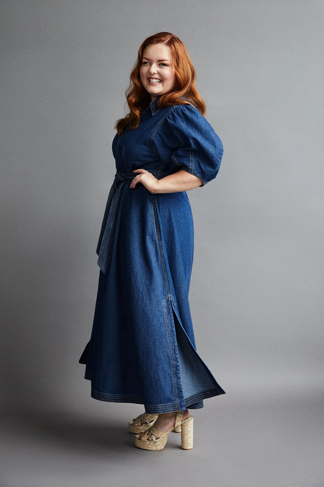 Lucy wearing a long denim dress and cork heeled sandals, standing against a grey background.