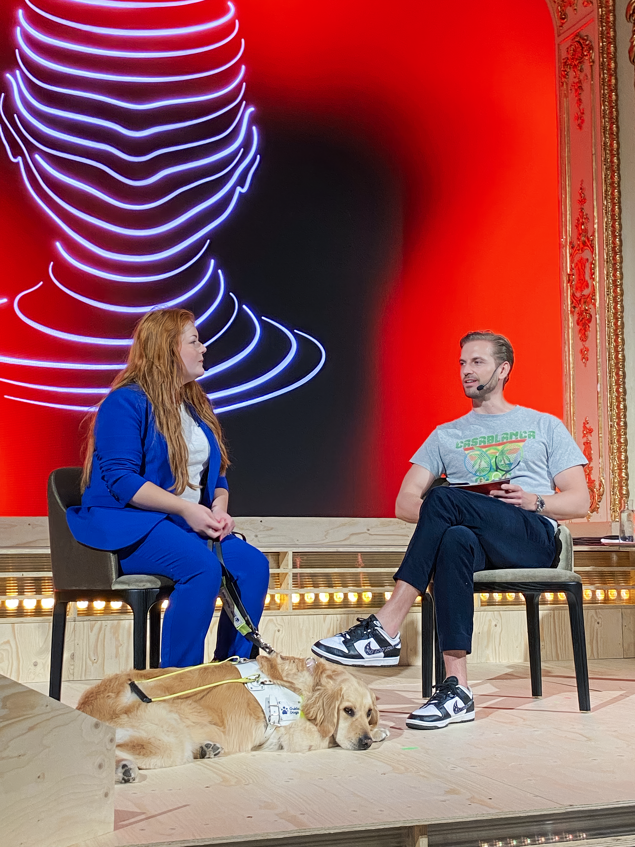 Lucy and Gustav sit on stage with a large neon art piece and ornate decorative frame behind them. Lucy on the left has long red hair and is wearing a blue blazer, while Gustav on the right is wearing a grey T-shirt, black pants, and sneakers.