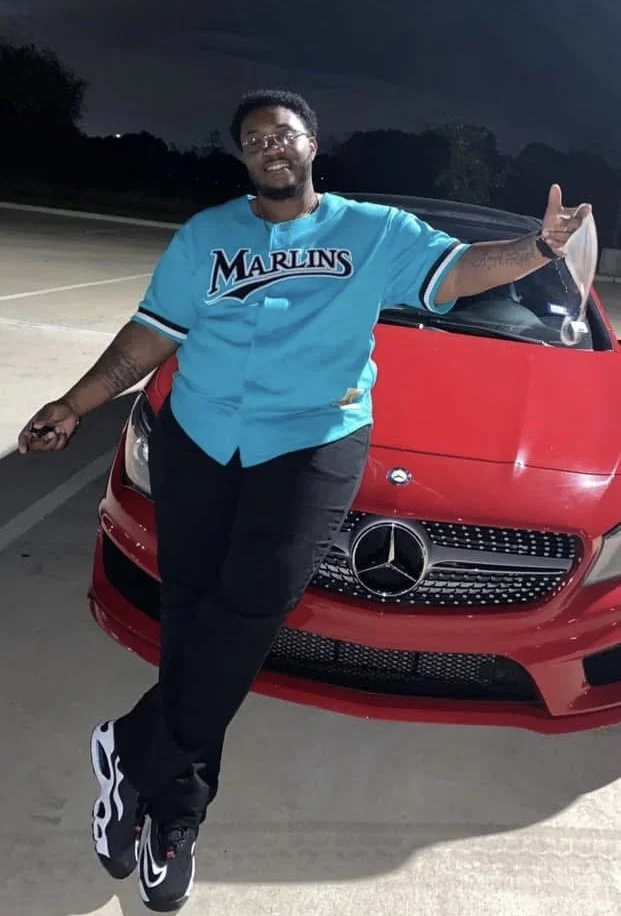 A man smiling and posing in front of a red Mercedes-Benz car at night, wearing a Miami Marlins jersey and holding an object in his right hand.