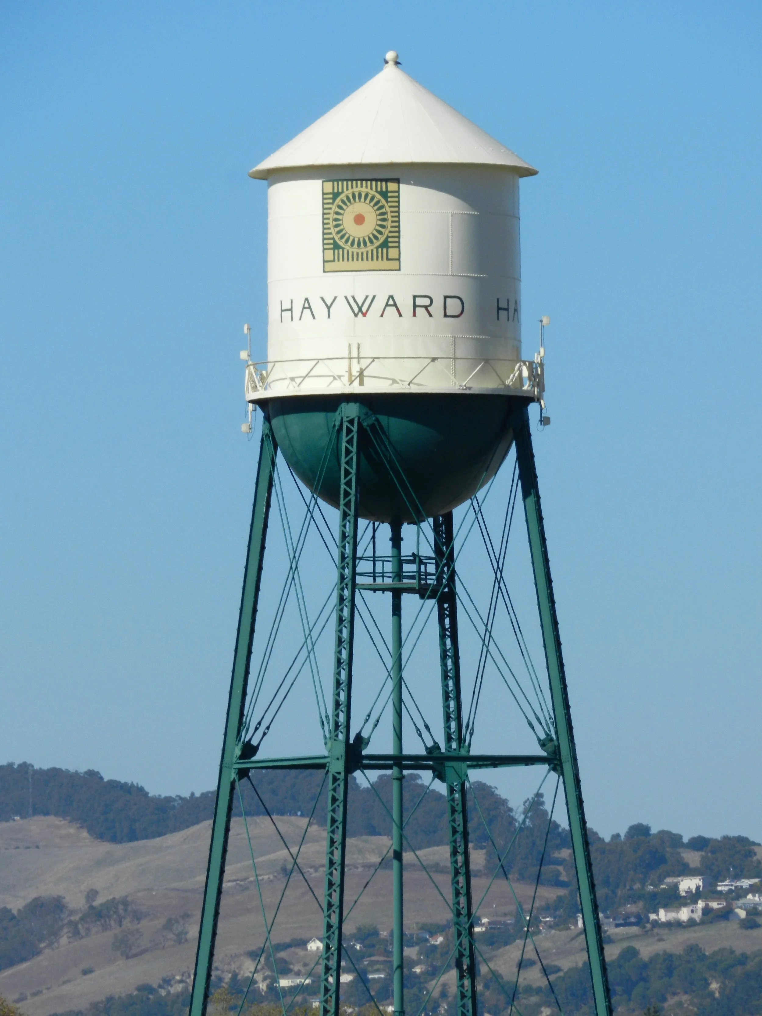 A water tower with a white conical roof, displaying the words "HAYWARD" on the side, set against a clear blue sky with distant rolling hills and some buildings below.