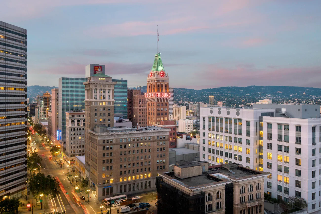 Cityscape of downtown Oakland, California at dusk, with illuminated buildings and streets, featuring the Tribune Tower with a clock tower and green spire.