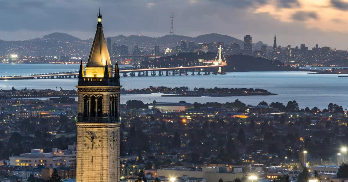 City skyline view of San Francisco at dusk with a prominent clock tower in Berkeley, CA, in the foreground and the Bay Bridge illuminated in the background.