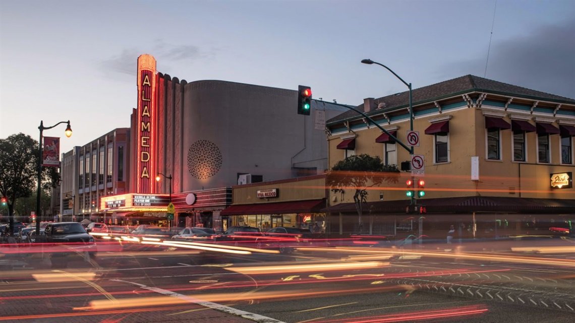 The Alameida theater on a city street during dusk, with streaks of car lights, traffic signals, and a historic building with yellow walls and purple awnings.