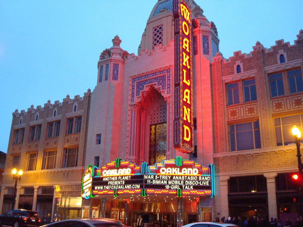 The Fox Oakland theater with a neon marquee advertising live music events, surrounded by parked cars and people outside, during dusk.