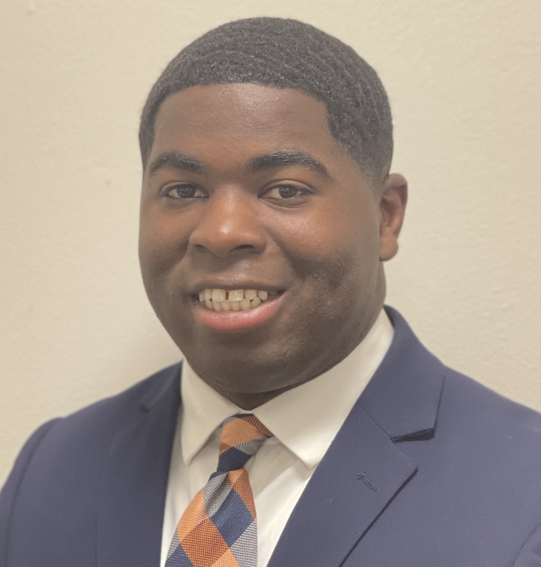 Photo of a smiling young man with short, textured hair, wearing a navy blazer, white shirt, patterned tie, and standing against a plain light-colored background.