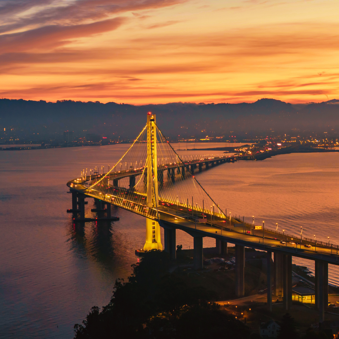 A bridge illuminated at sunset over a body of water, with a dramatic sky filled with orange and purple hues, and city lights in the distance.
