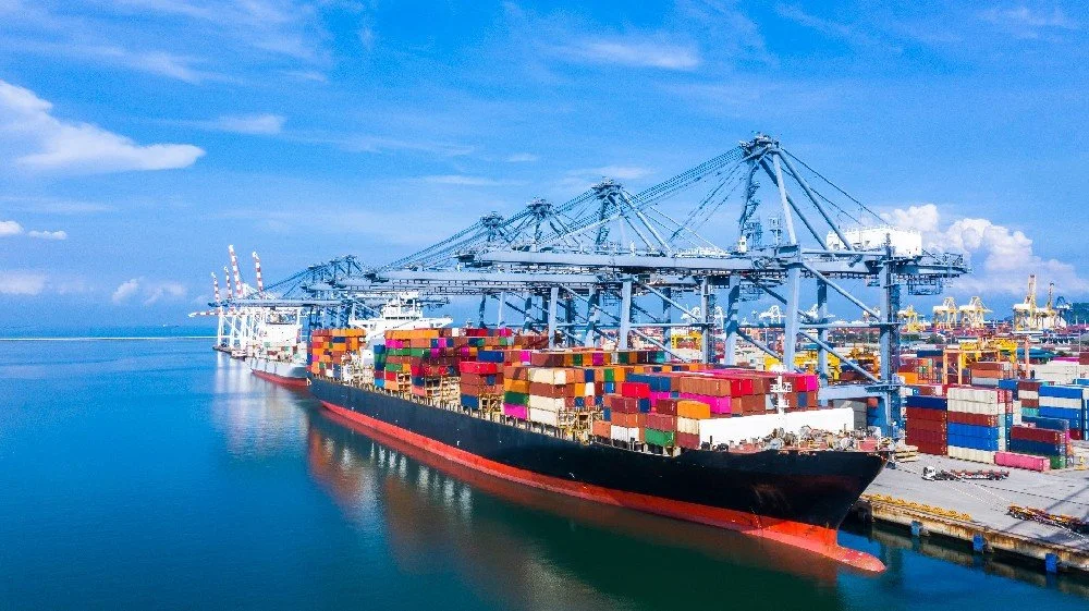 Large cargo ship docked at a port with numerous colorful shipping containers and blue gantry cranes under a partly cloudy sky.