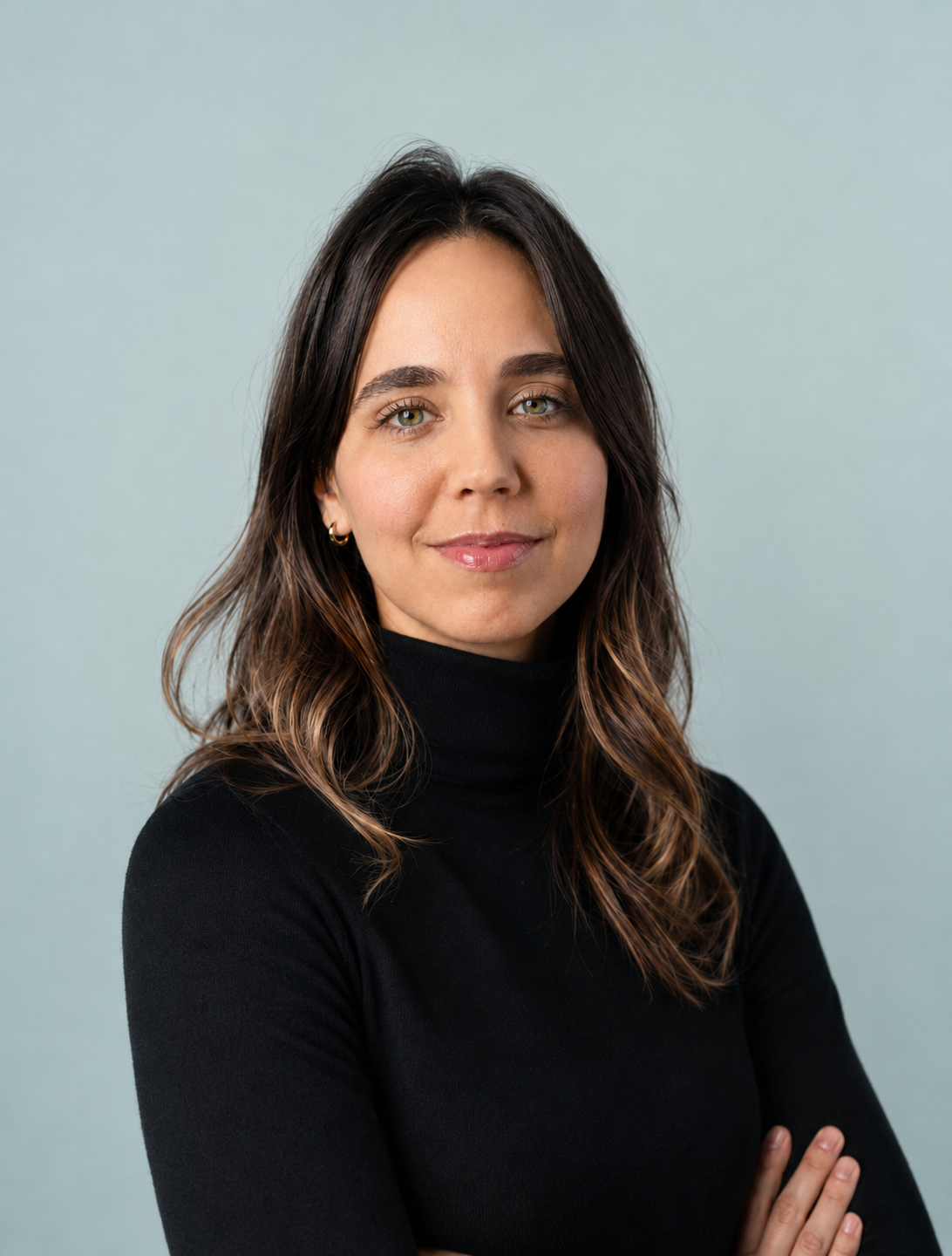 Portrait d'une femme aux cheveux bruns ondulés portant un pull noir, souriant légèrement, avec un fond gris clair.