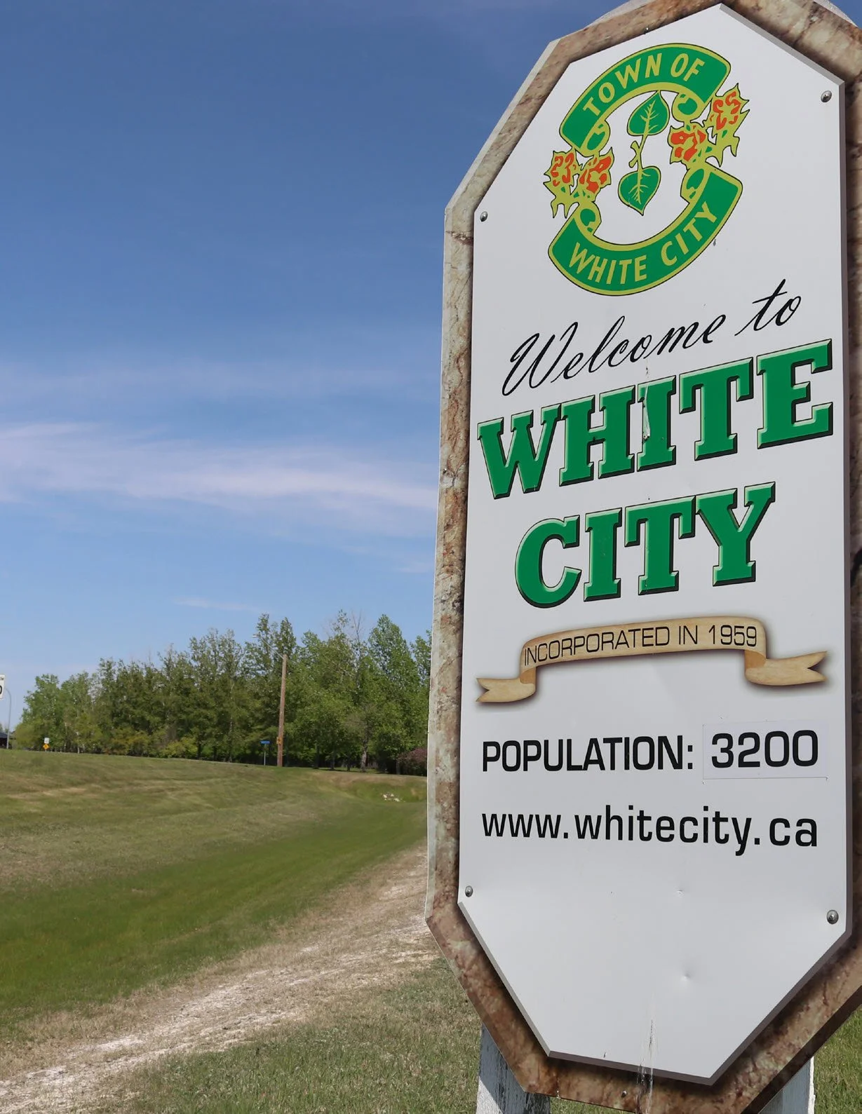 Sign welcoming visitors to White City, featuring green and yellow emblem, with additional details about foundation in 1959, a population of 3,200, and the website www.whitecity.ca. The sign is outdoors with greenery, trees, and a partly cloudy sky in the background.