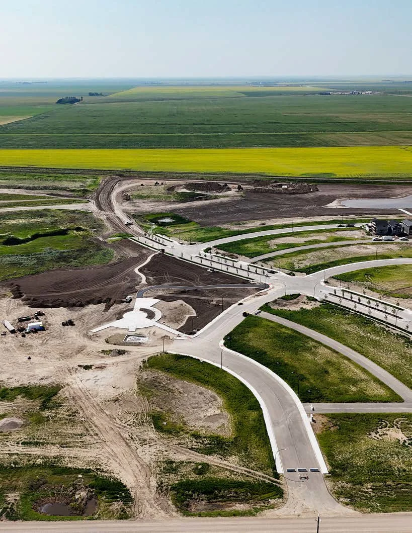 Aerial view of a construction site with new roads, some completed and some under construction; surrounding farmland and fields in the background.