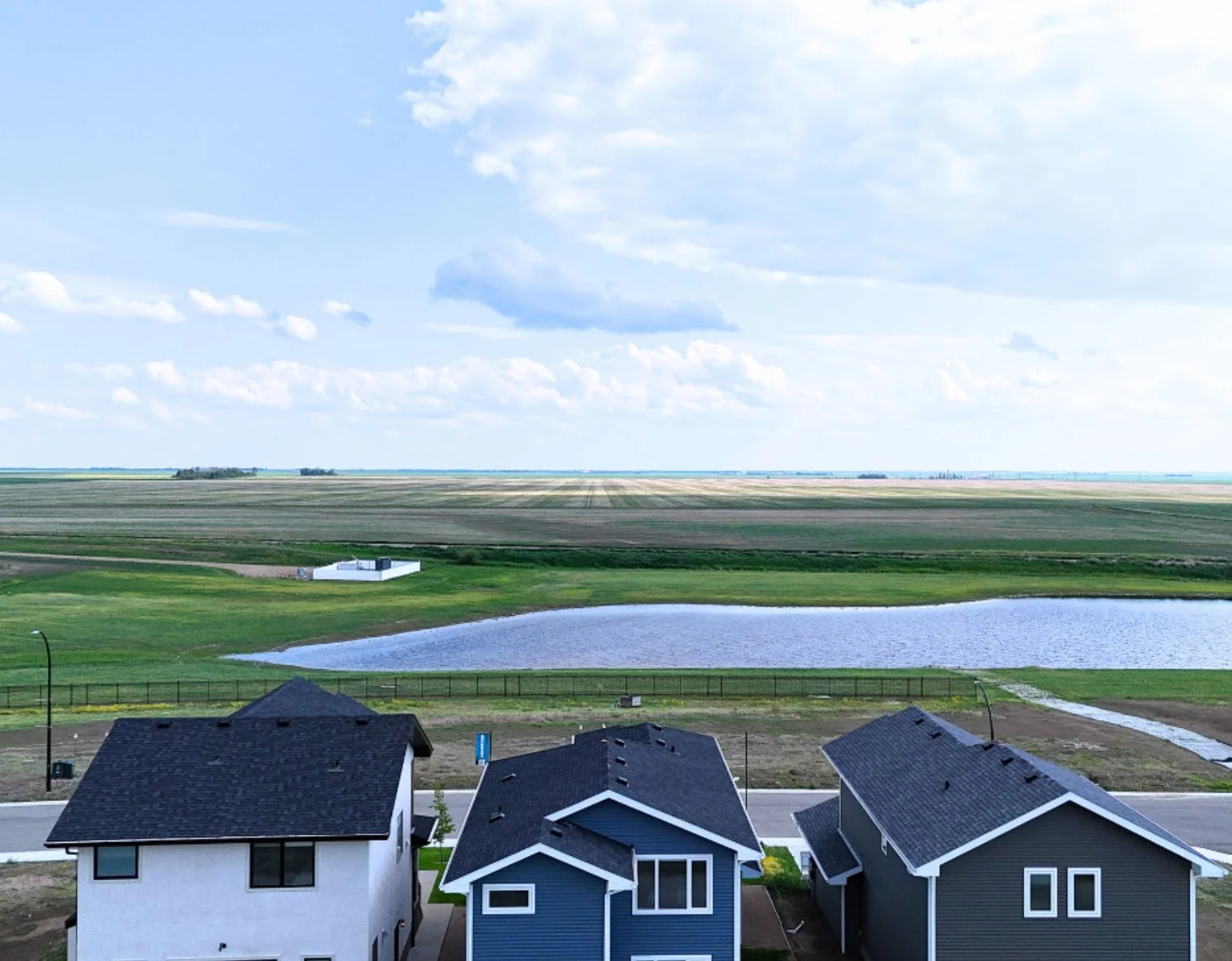 A suburban neighborhood with modern houses in the foreground, a pond, and expansive fields and farmland stretching to the horizon under partly cloudy skies.