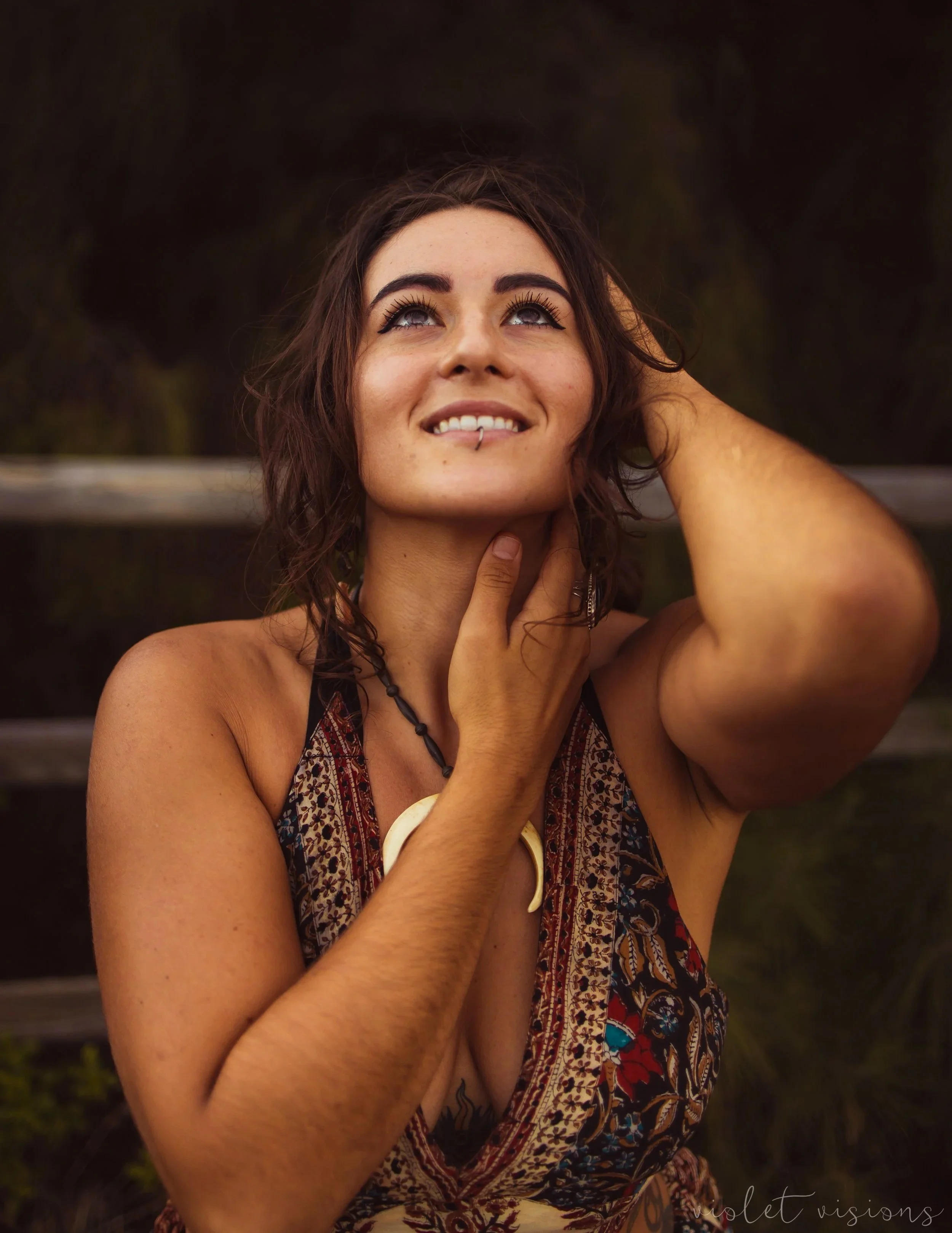 A young woman with dark brown, wavy hair, blue eyes, and makeup, smiling softly with her hand on her neck, wearing a patterned sleeveless dress and accessories, outdoors against a blurred dark natural background.