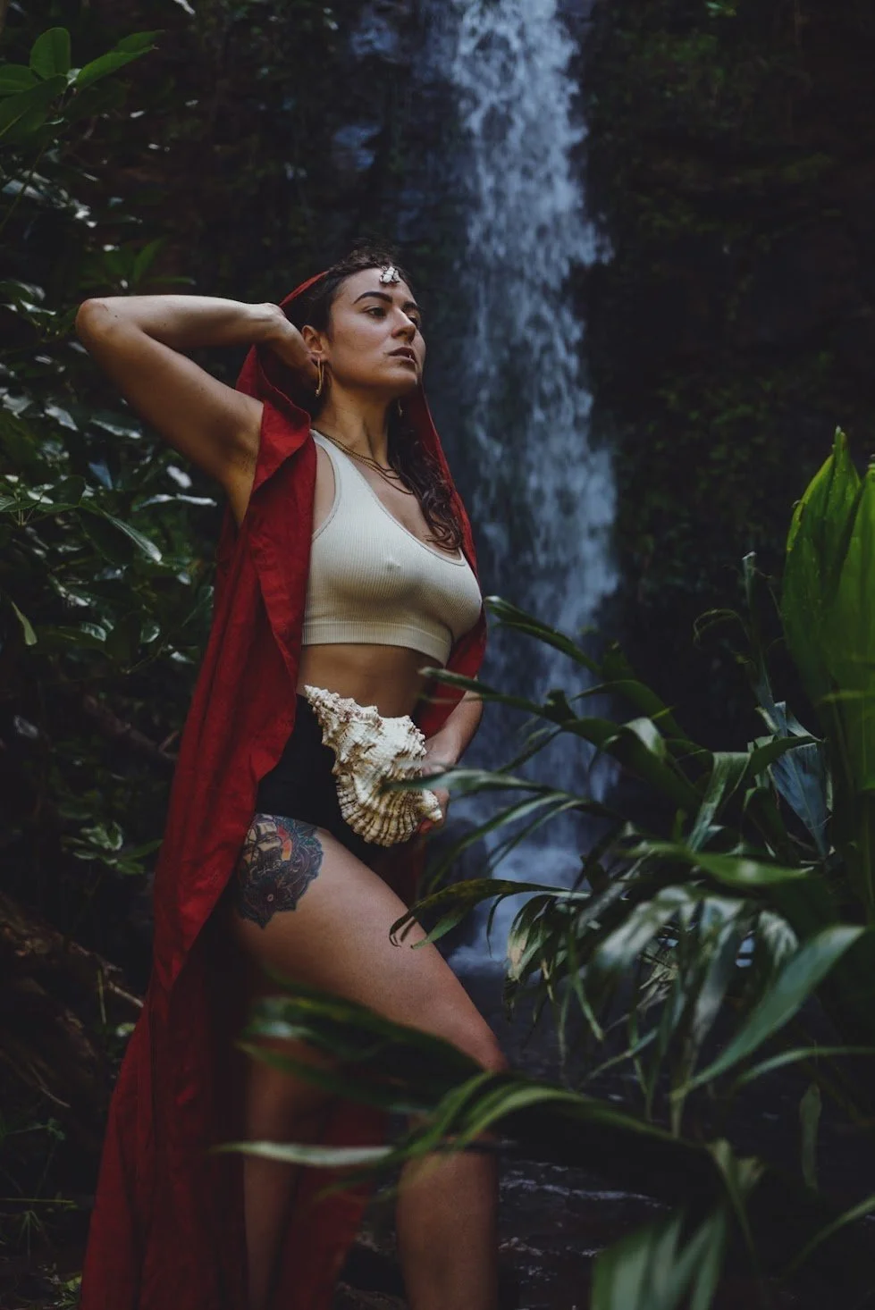A woman standing in front of a waterfall surrounded by lush greenery, wearing a cream crop top, black shorts, a red cloak, holding a seashell, with a tattoo on her thigh, and jewelry on her head and ears.