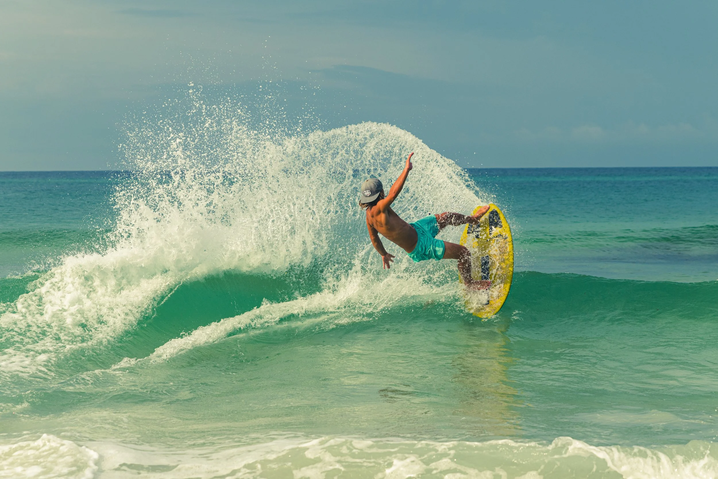 A shirtless man wearing teal shorts and a gray cap surfing on a yellow skimboard in the ocean, creating a splash as he rides a wave under a blue sky.